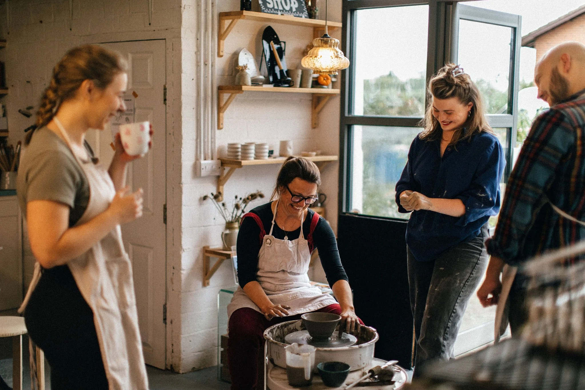 People engaging in a pottery class inside a studio, with a woman shaping clay on a potter's wheel, others observing, and a woman enjoying a hot drink.