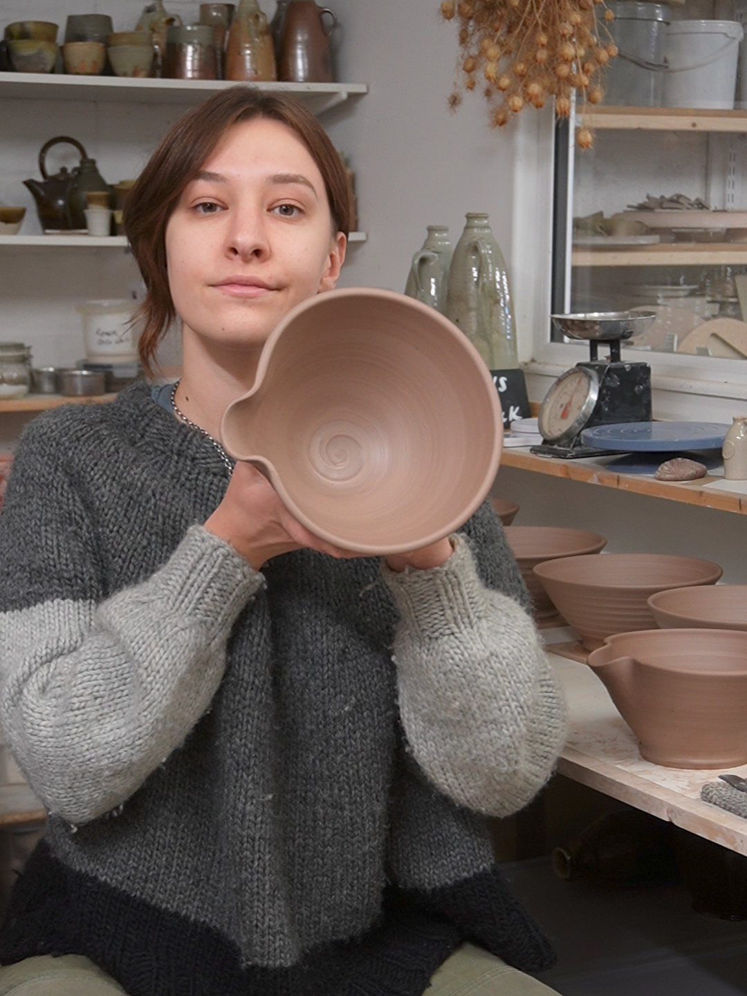 A woman standing in a pottery studio holding an unglazed ceramic bowl. There are completed bowls and shelves with pottery in the background.
