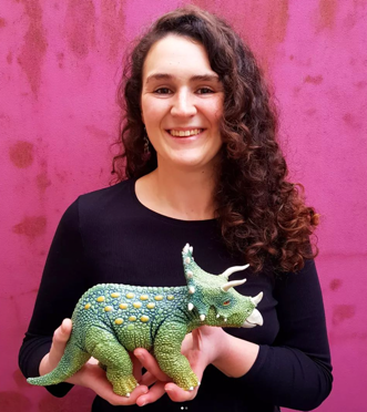 Woman with curly hair smiling, holding a detailed green and yellow ceramic triceratops dinosaur, standing in front of a pink background.
