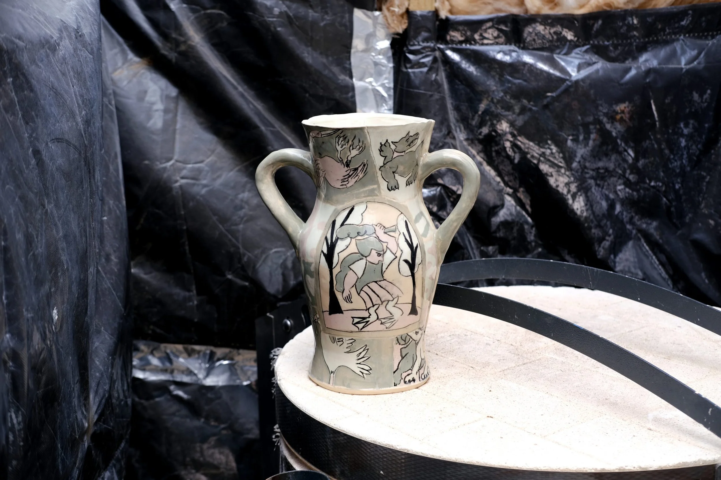 Ceramic vase with decorative sketches of animals and a woman, placed outdoors on a white circular surface with black plastic material in the background.