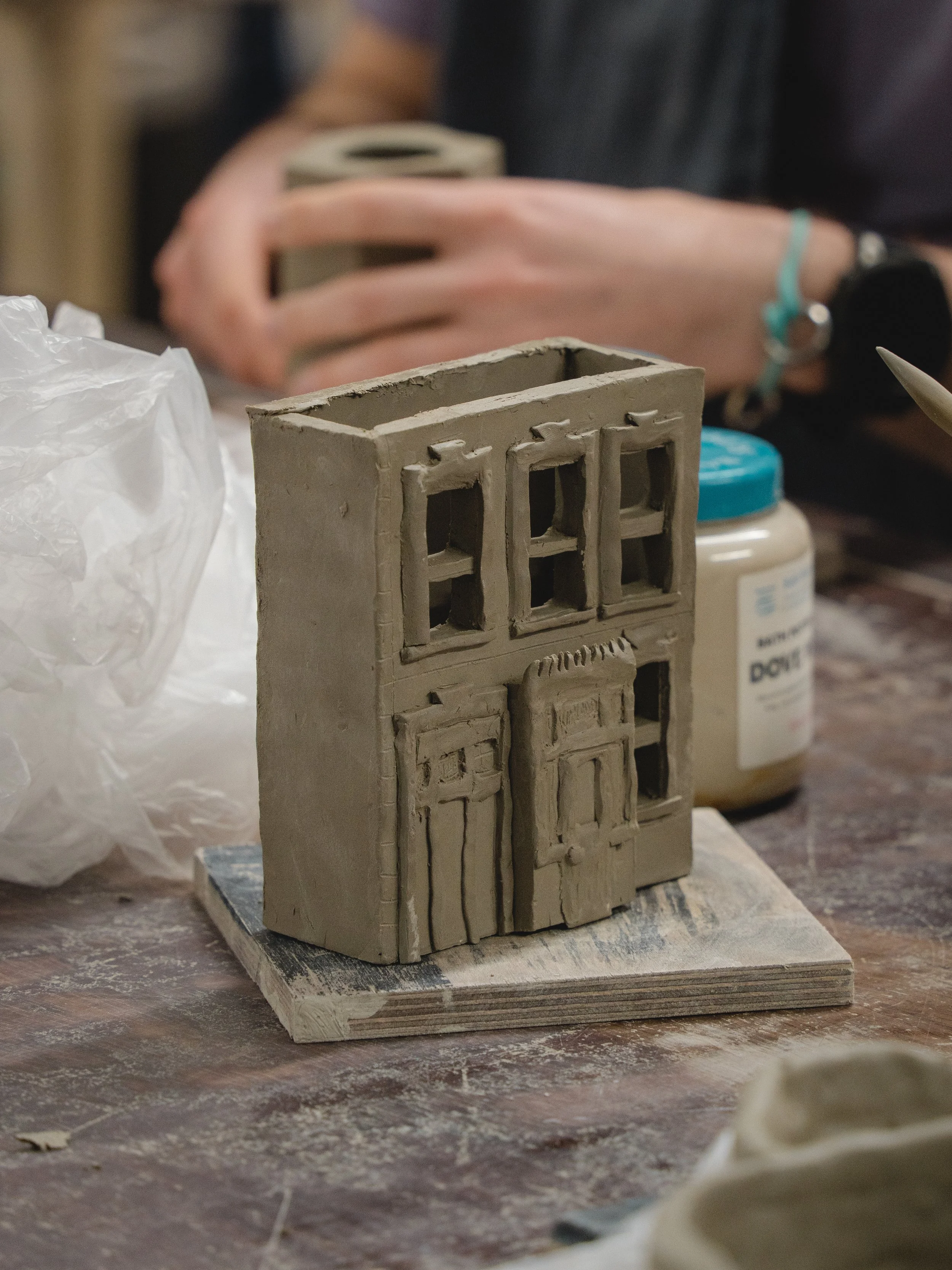 An unfinished miniature clay building model with windows and doors, placed on a wooden board. In the background, a hand is holding a small piece of clay, surrounded by art supplies and a white container.