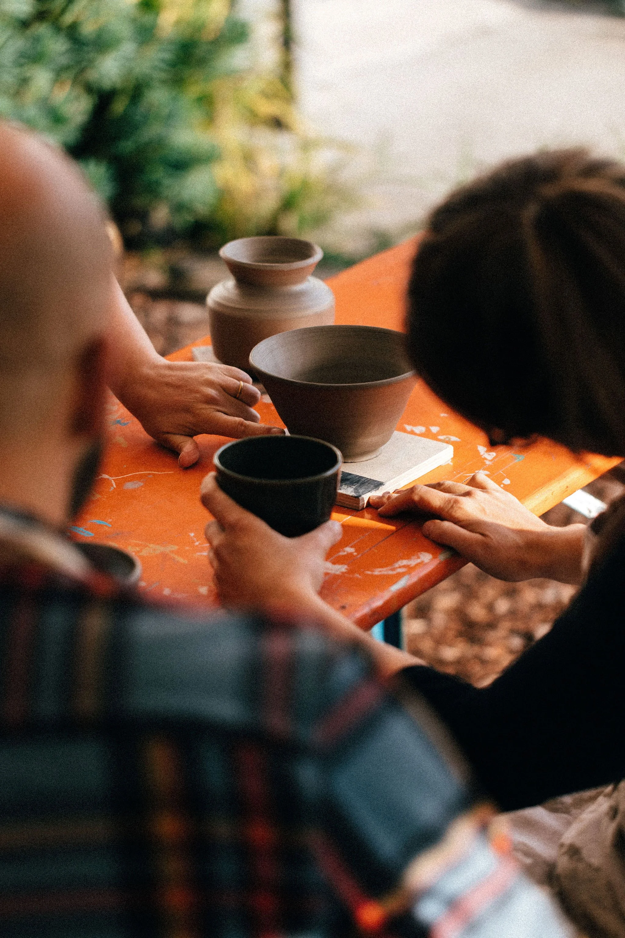 People gathered around an orange table, looking at pottery and holding cups, with a pottery vase on the table.