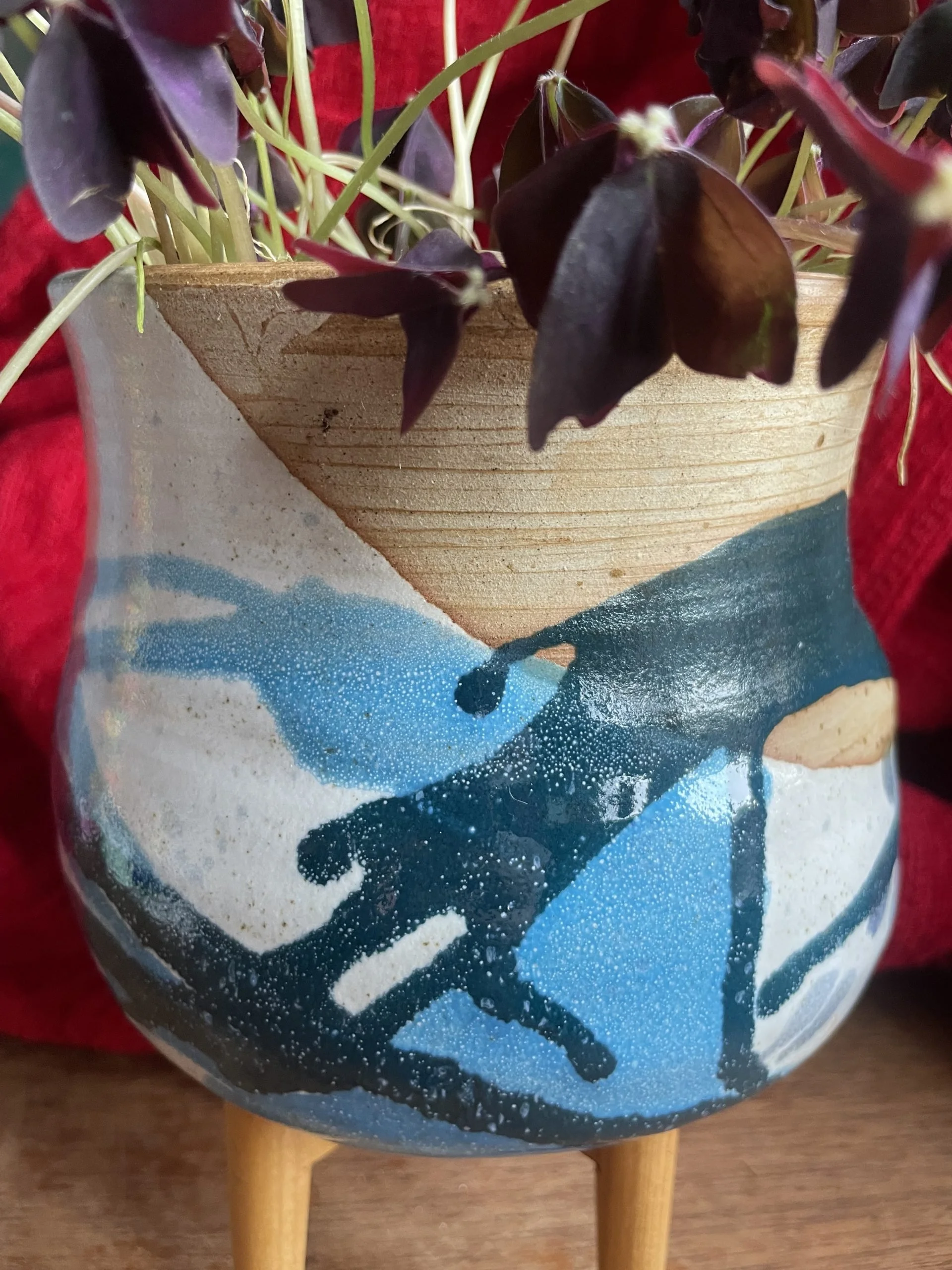 A round blue and white ceramic vase with legs, with a beach scene painted on it, showing waves, sand, and a dolphin, with a potted plant with dark purple leaves placed in it.