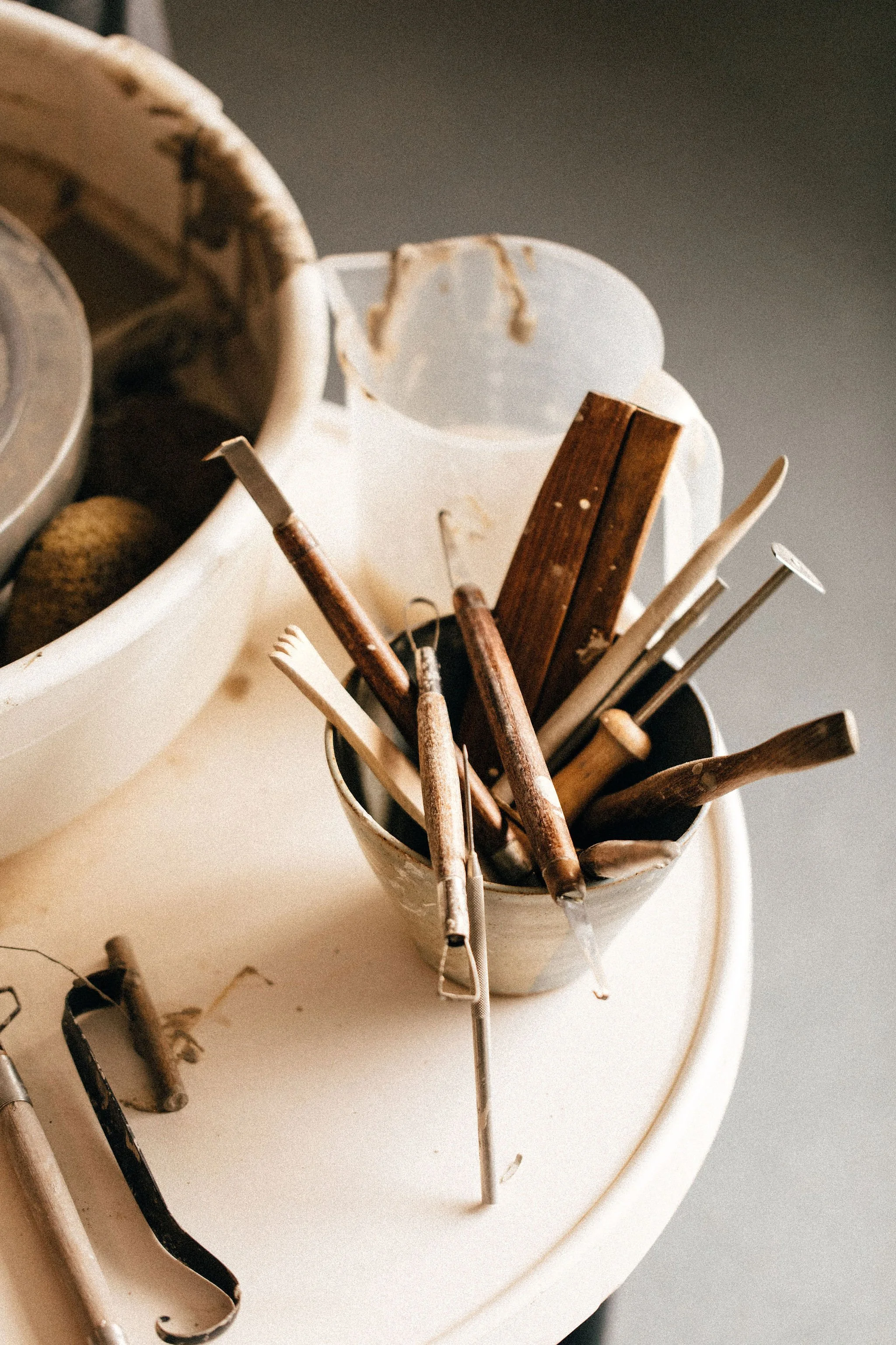 A collection of ceramic sculpting tools in a small bowl, placed on a beige surface, next to a pottery throwing wheel