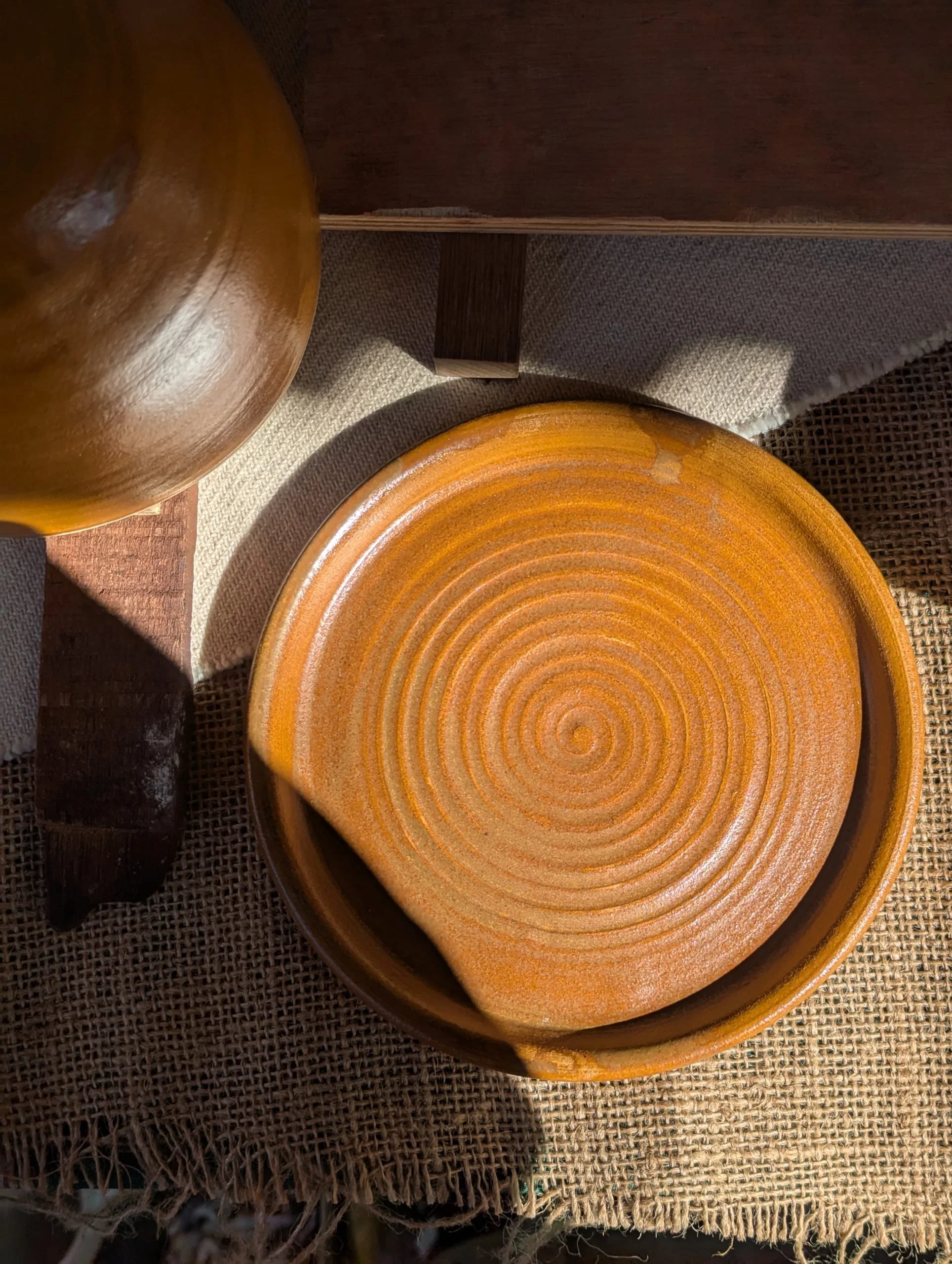 Top-down view of a brown glazed ceramic plate with concentric circle patterns inside, placed on a woven fabric surface. Part of another brown ceramic object is visible on the top left corner.