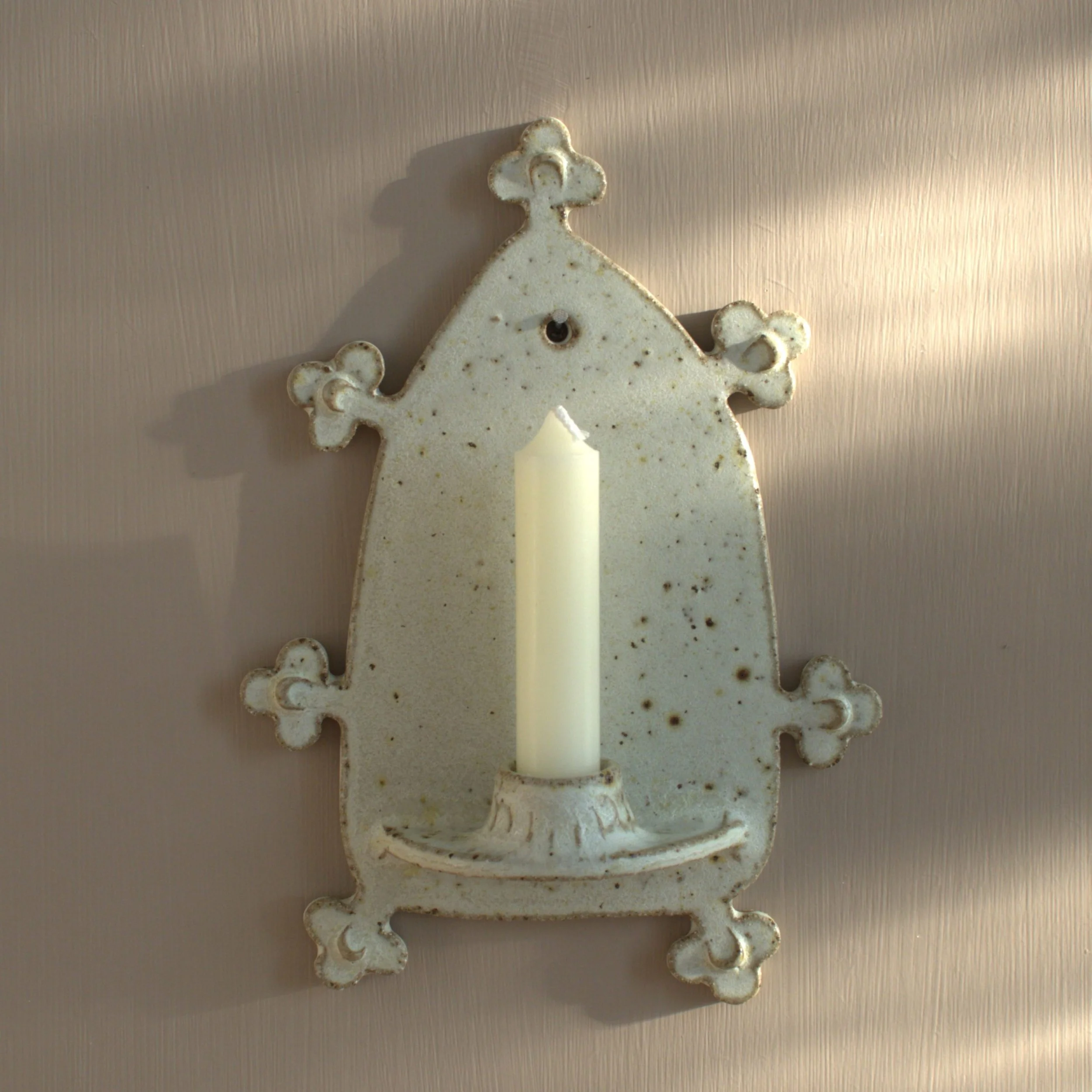 A beige ceramic wall sconce ornament shaped like an arch, placed on a light-colored surface. The ornament has a small hole to hang it, with a tall white candle held at the bottom in the candle holder.