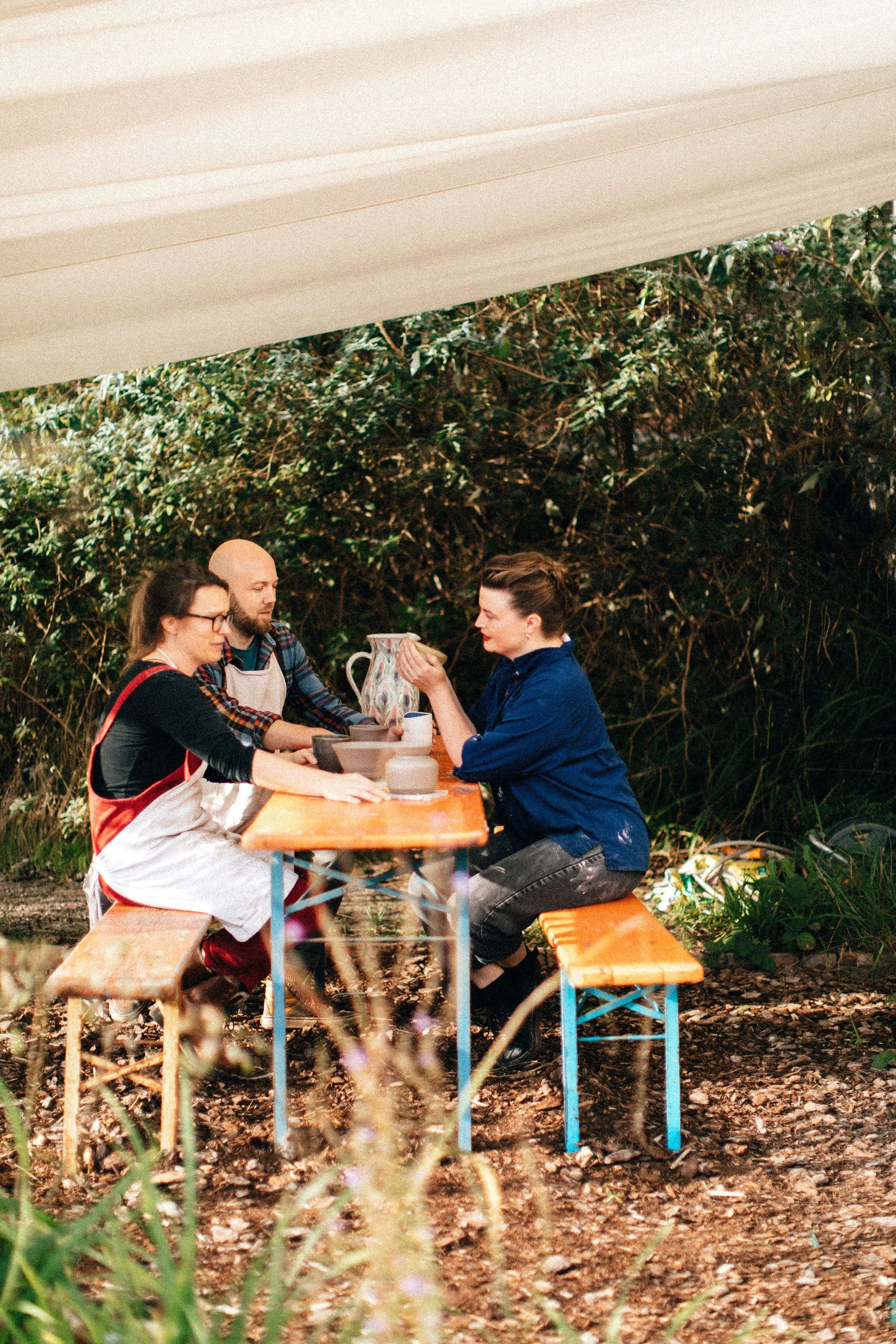 Three people sitting at a wooden picnic table outdoors under a canopy, with greenery in the background, engaged in conversation.