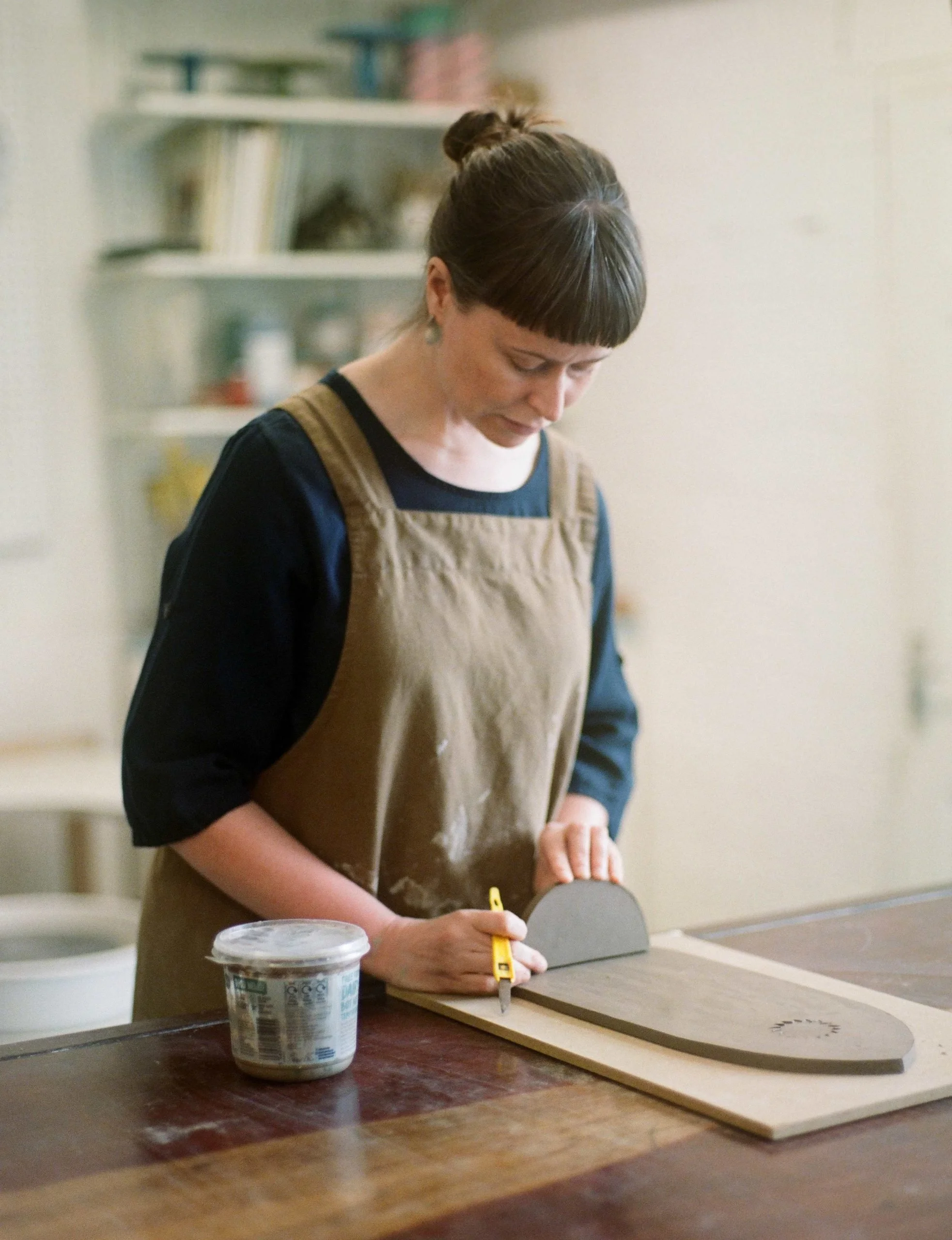 A woman in a brown apron and dark shirt working on flat clay project at a table, with a container of water nearby.
