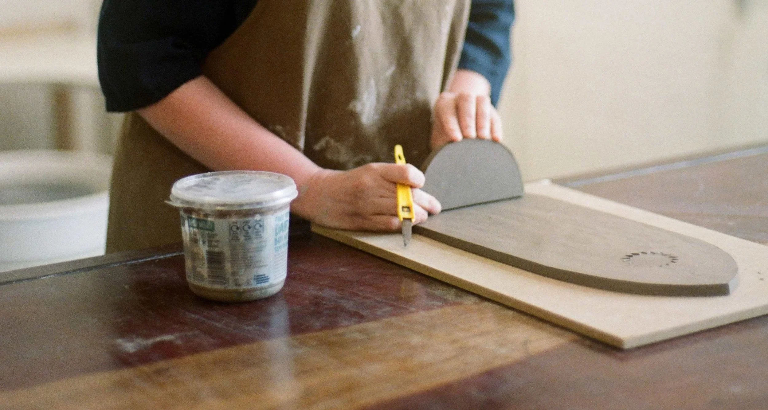 Person wearing a brown apron and black shirt using a utility knife to cut a piece of clay material on a work table. A container of water is placed on the table near the person.