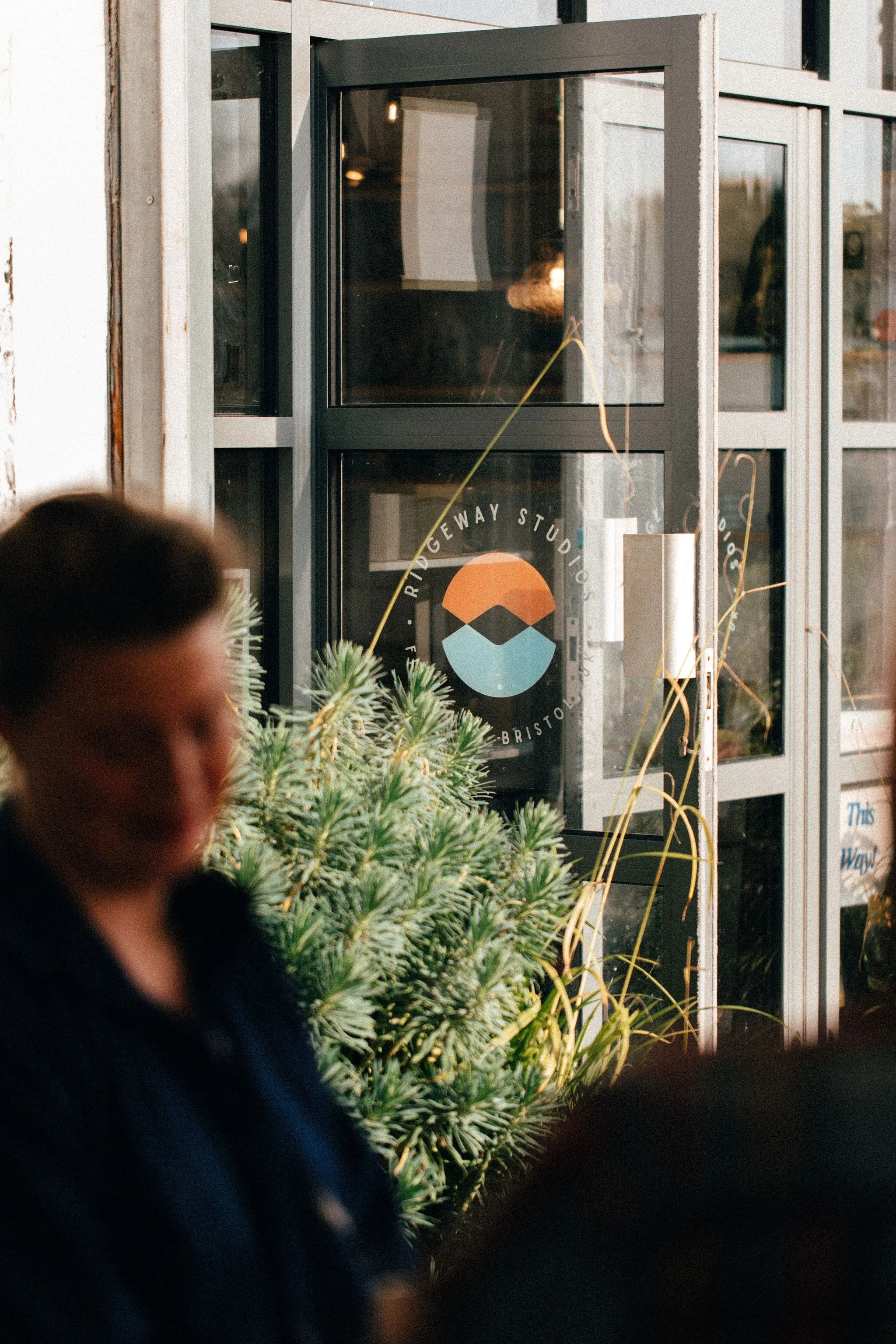 A storefront window with a logo and text, partially obscured by plants in the foreground. The logo consists of a stylized mountain with a sun or moon above it. A person with brown hair is seen in the foreground, partially out of focus.