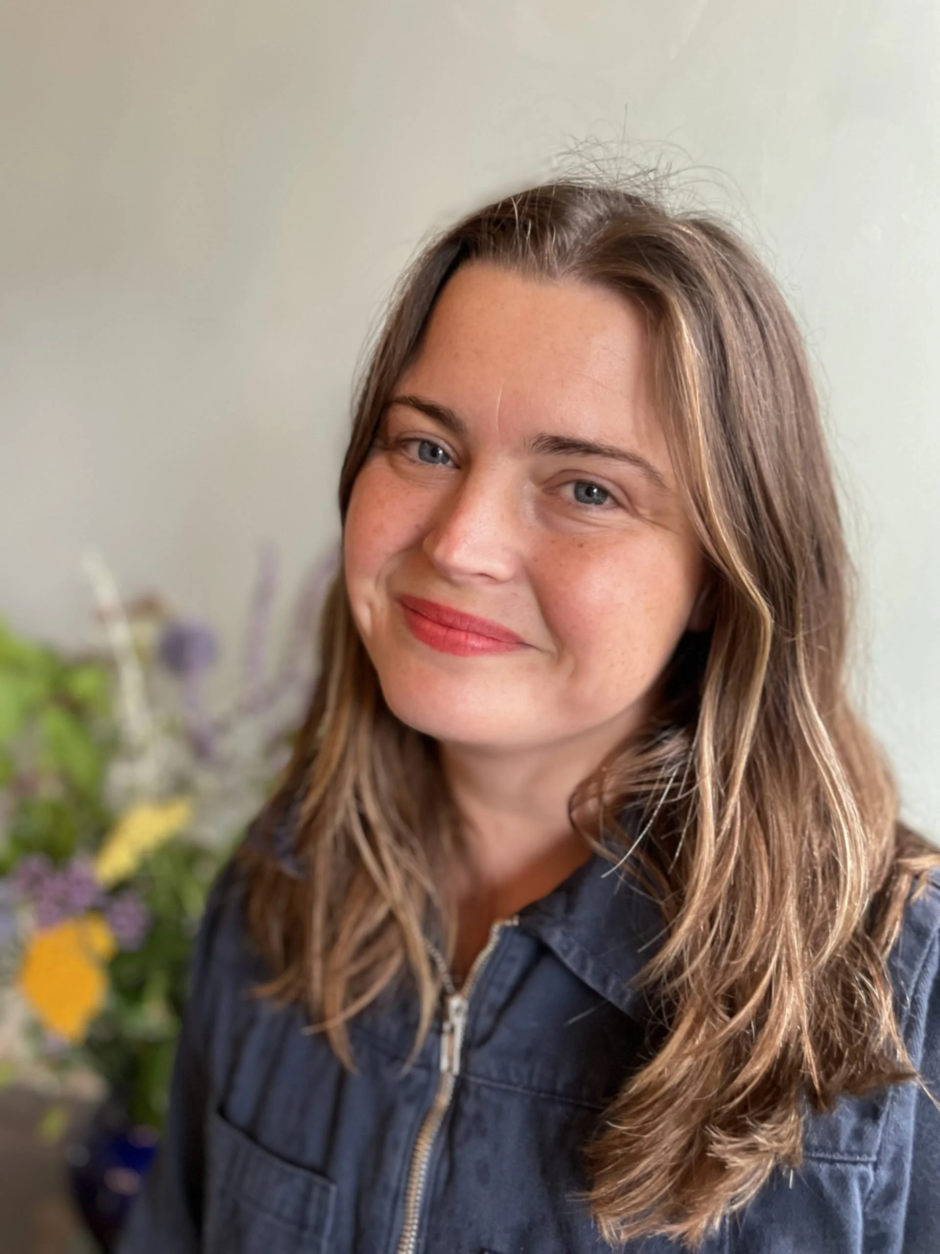 A young woman with long, wavy brown hair and blue eyes smiling in front of a blurred background of colorful flowers, wearing a dark jacket with a zipper.