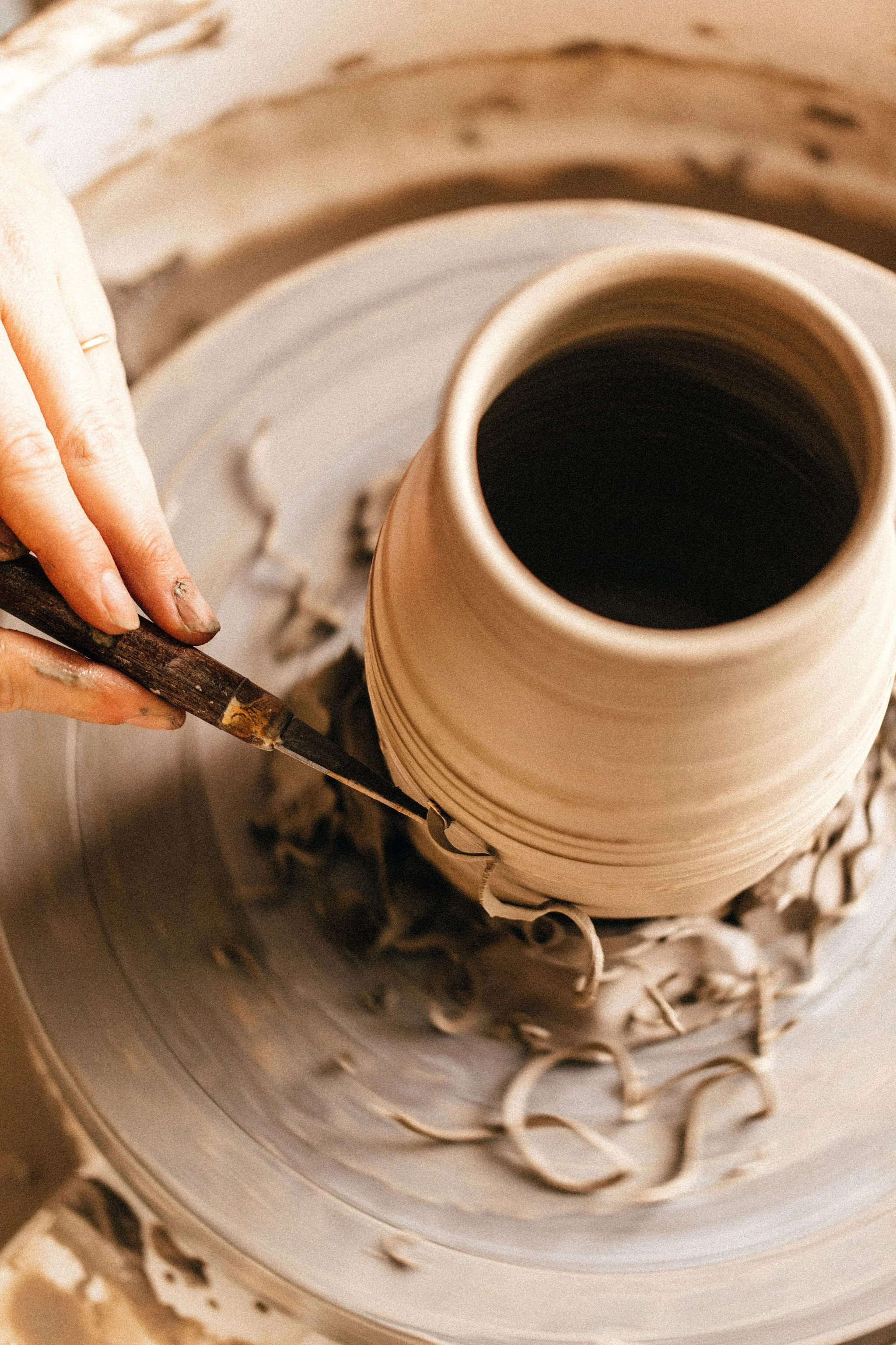 A person shaping a clay pot on a pottery wheel, with their fingers pressing on the clay as it spins. They are trimming the excess clay using a tool.