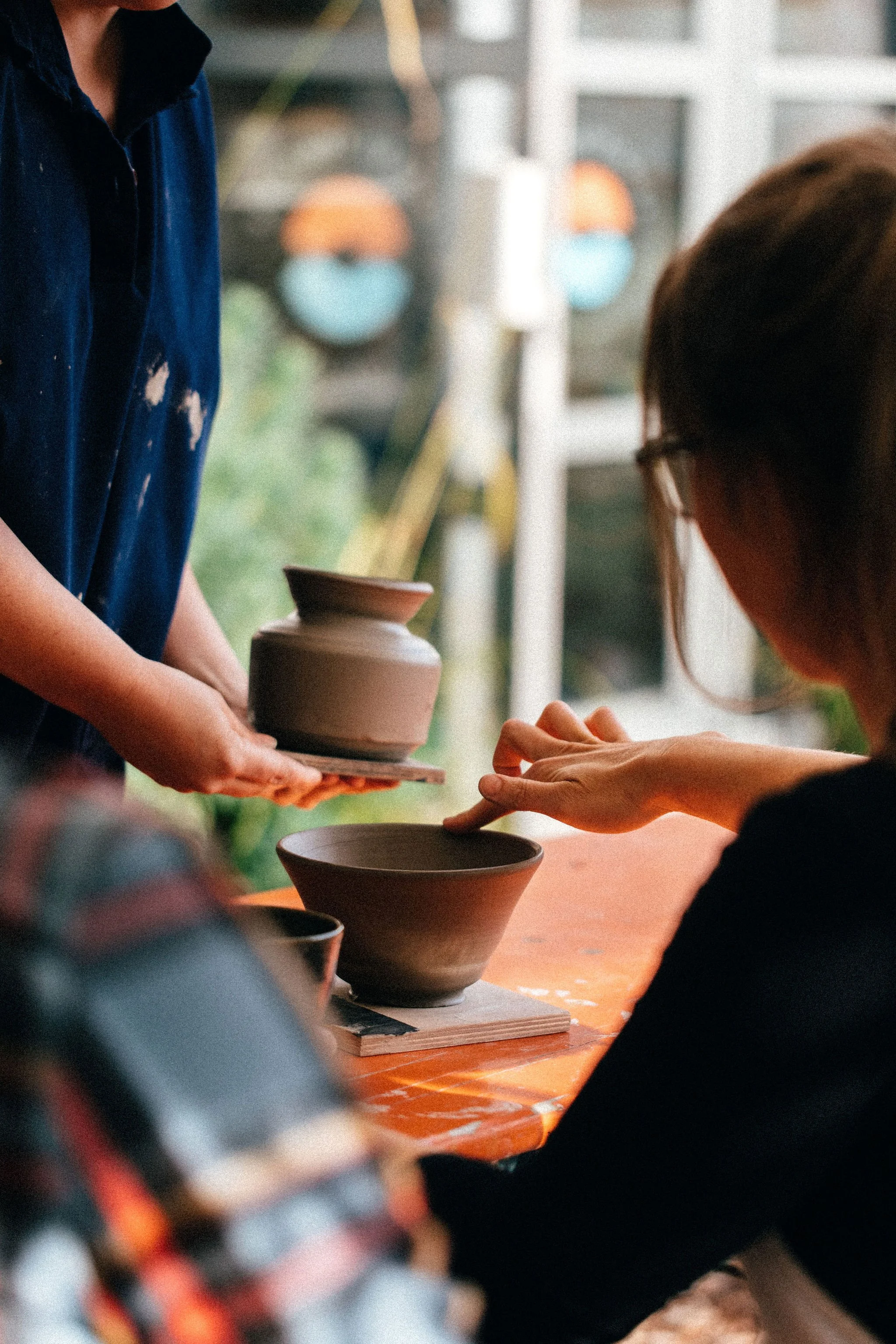 Person in dark shirt handing handcrafted ceramic bowl to another person with glasses at a pottery workshop.