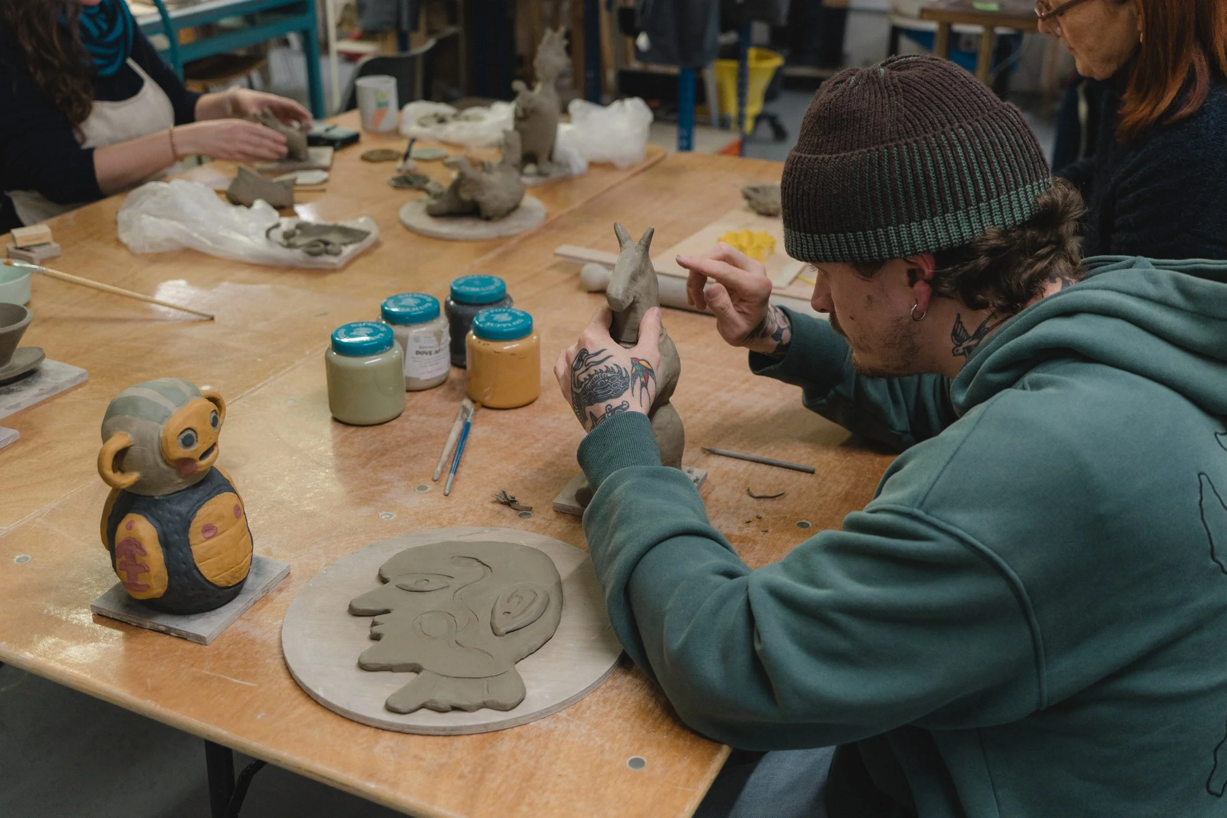 People working on pottery sculptures at a workshop table, including a person wearing a brown knit hat, sculpture of an animal with large ears, and various clay figures and tools spread across the table.