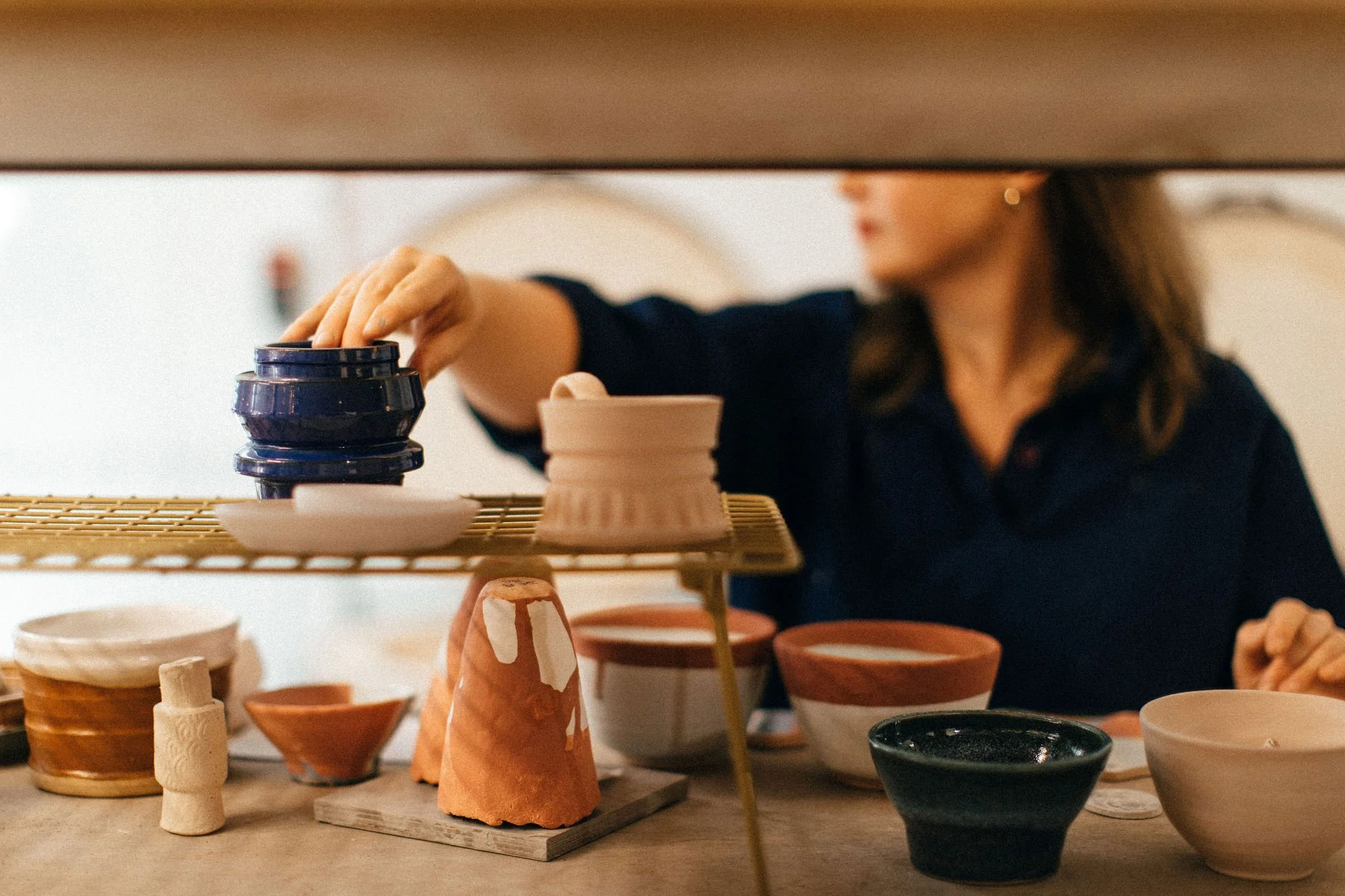 A woman arranging small pottery bowls on studio shelves, with various ceramic pieces on the shelves in front of her