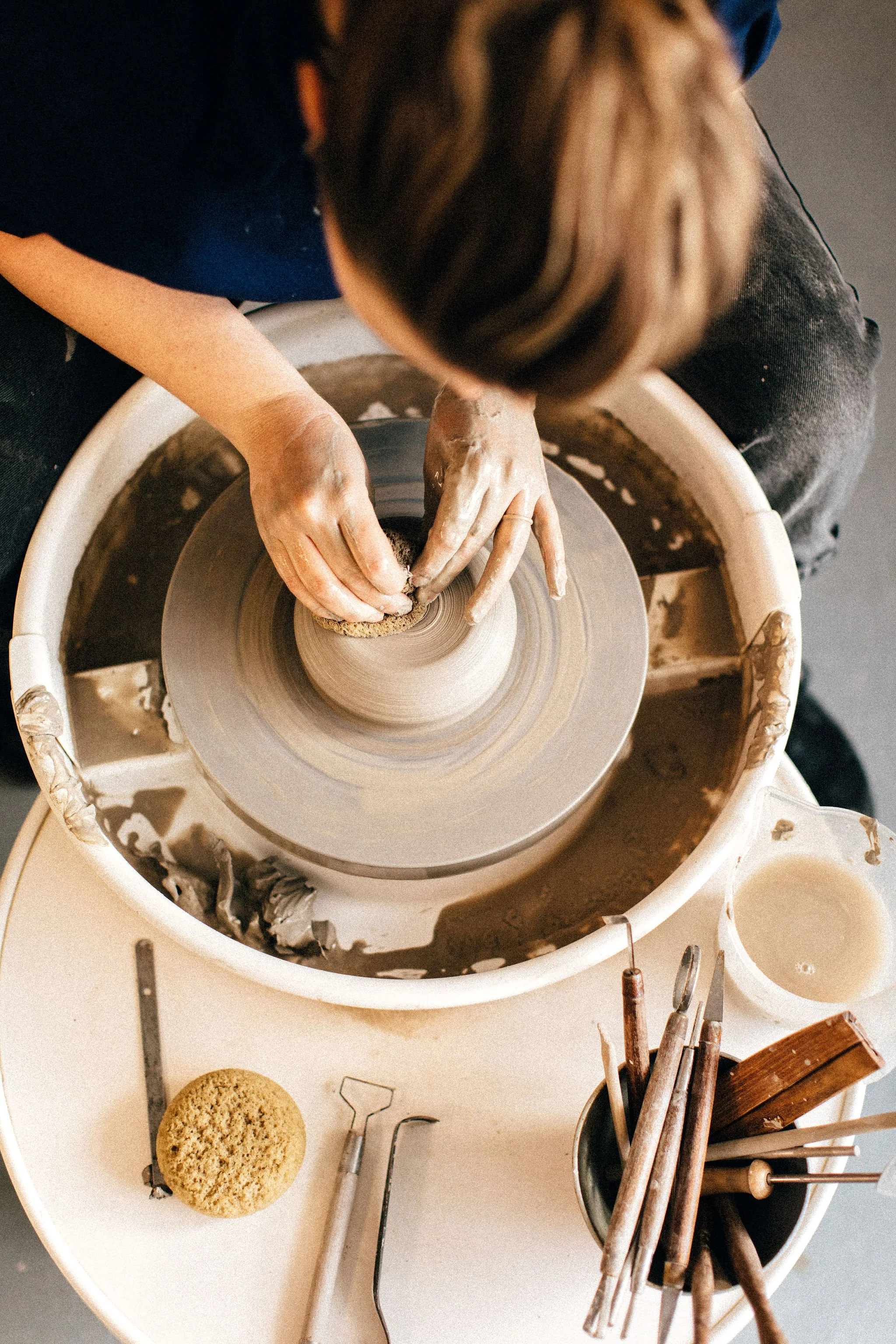 The image shows a person making pottery on a pottery wheel. Their hands are shaping the clay as the wheel spins. The workspace has various pottery tools and materials, including a sponge, metal tools, and clay. There is also a round sponge on the table.