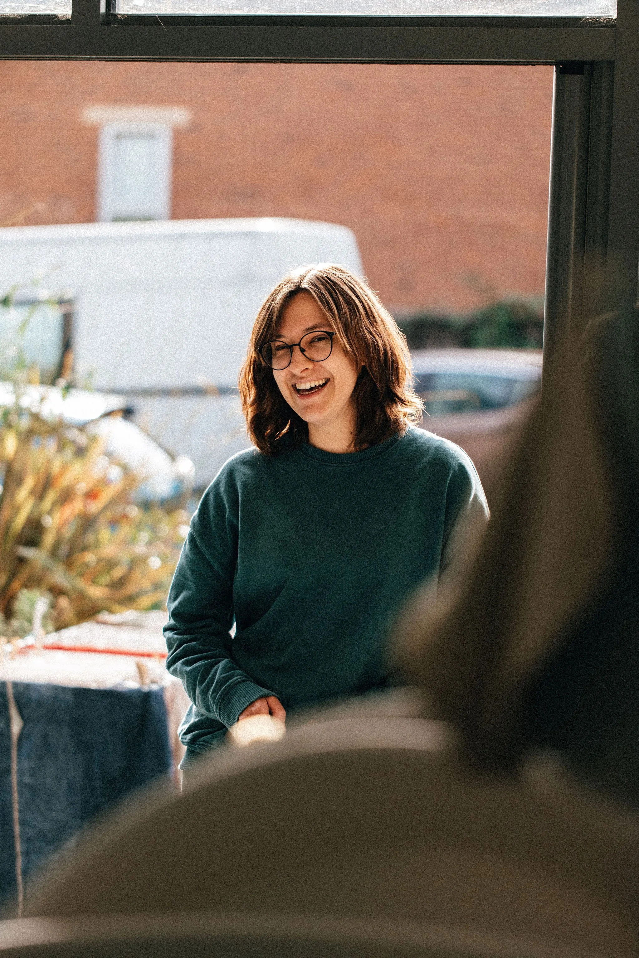 A woman with shoulder-length brown hair, glasses, and a dark sweater, smiling and looking to the side, indoors near a window with urban background.