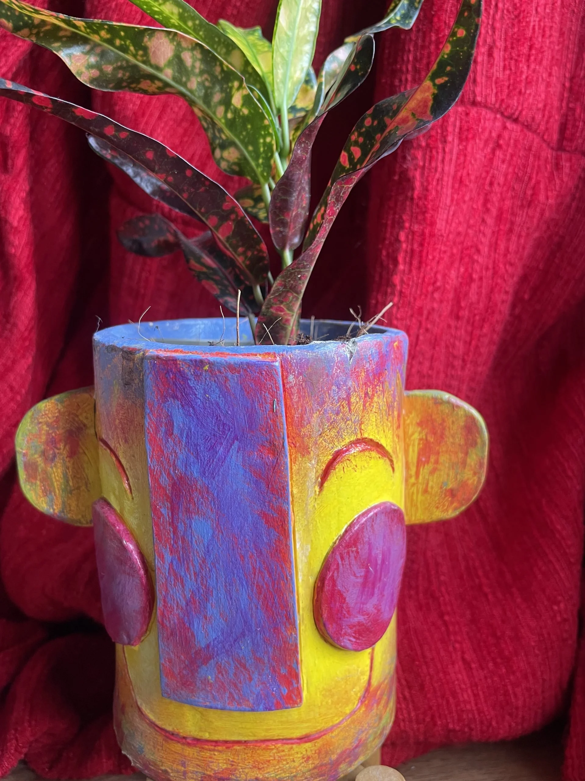 Colorful glazed vase with pink hearts, filled with a plant with green and red variegated leaves, held in front of a red textured background.