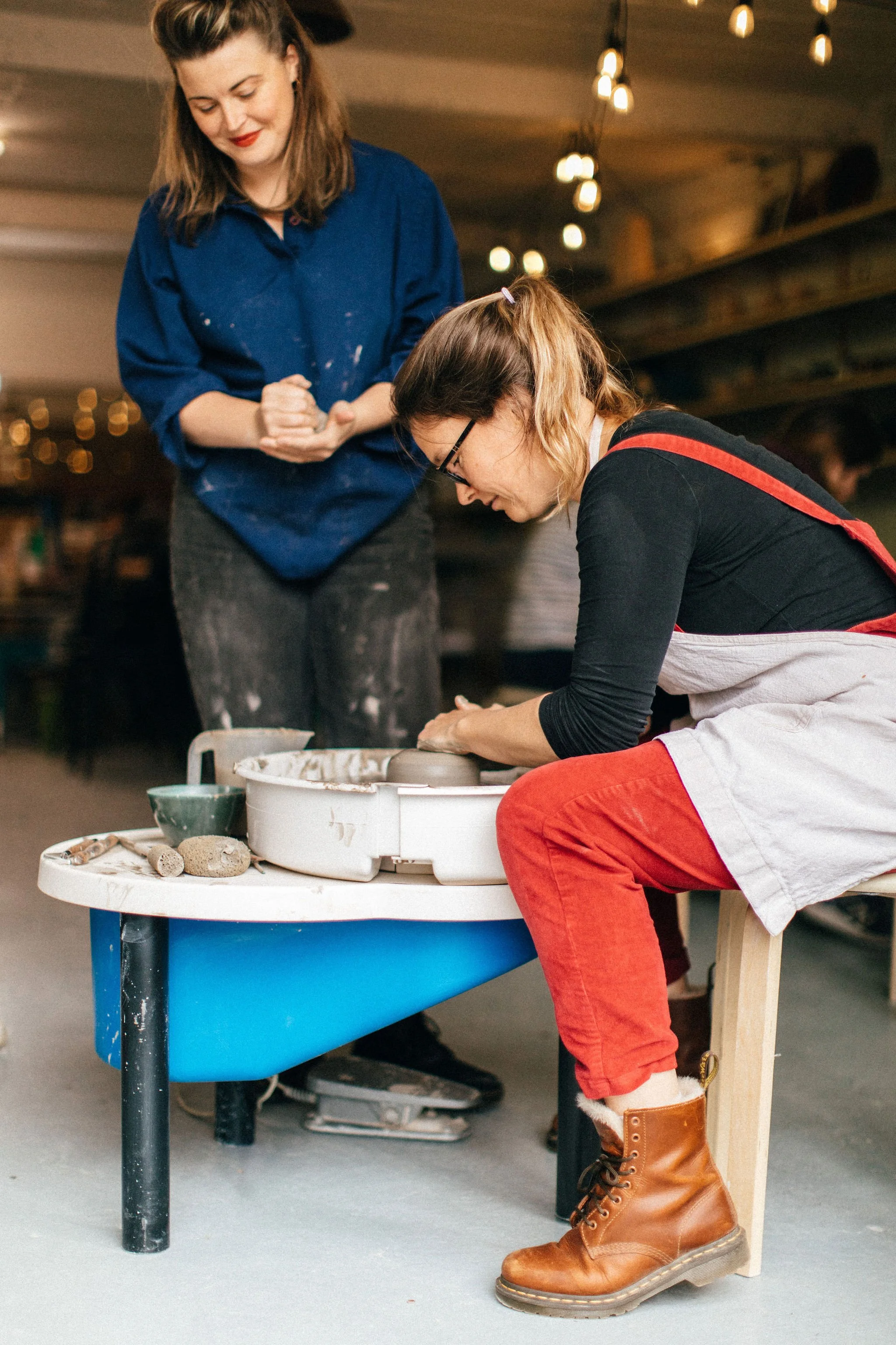 A woman wearing brown boots and red pants is sitting on a chair and working on a pottery wheel in a studio. Another woman, standing nearby, is observing and smiling. The studio is filled with warm lighting and has a wooden beam ceiling.