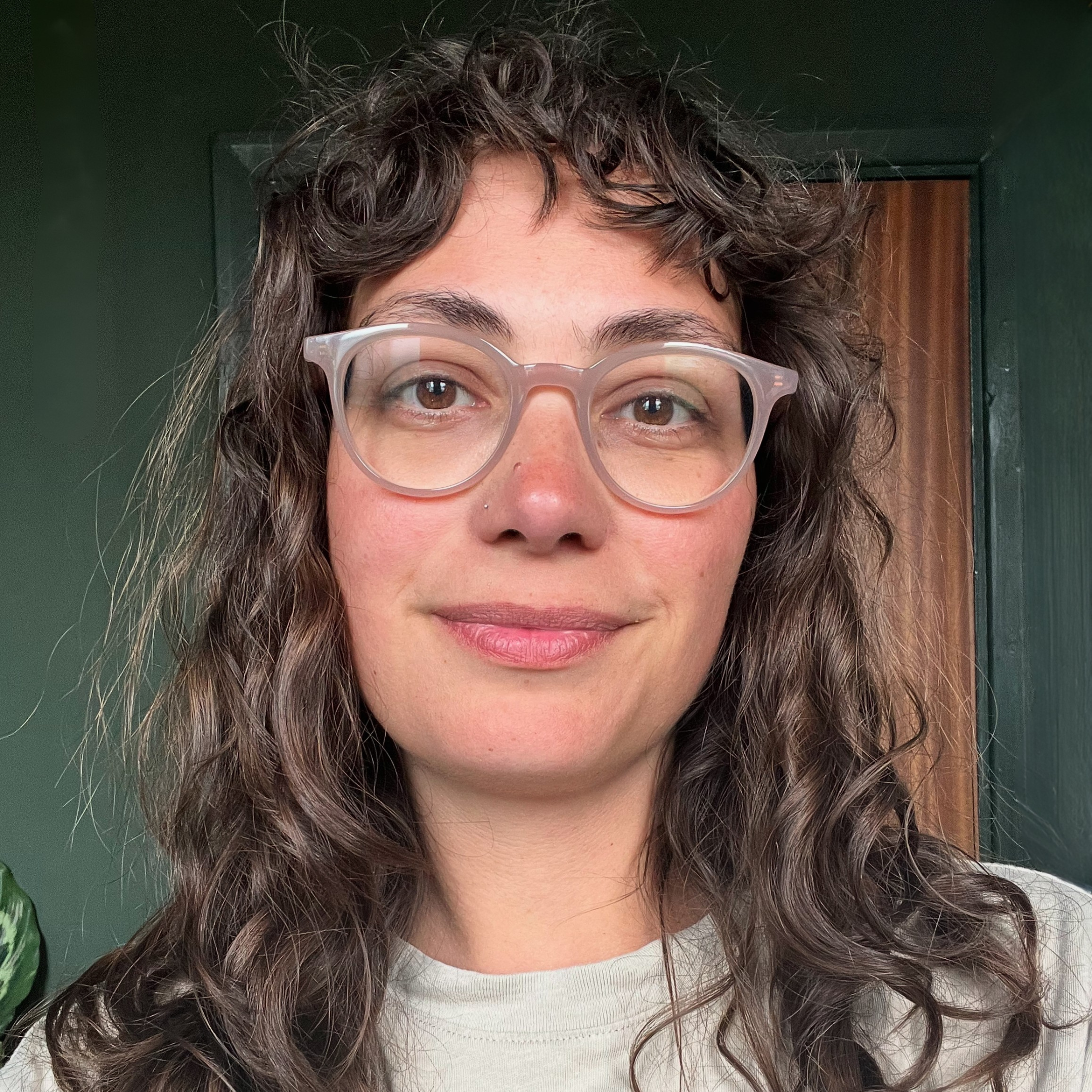 Close-up of a woman with curly brown hair and light skin wearing translucent glasses and a white top, standing in front of a dark green wall with a wooden door in the background.