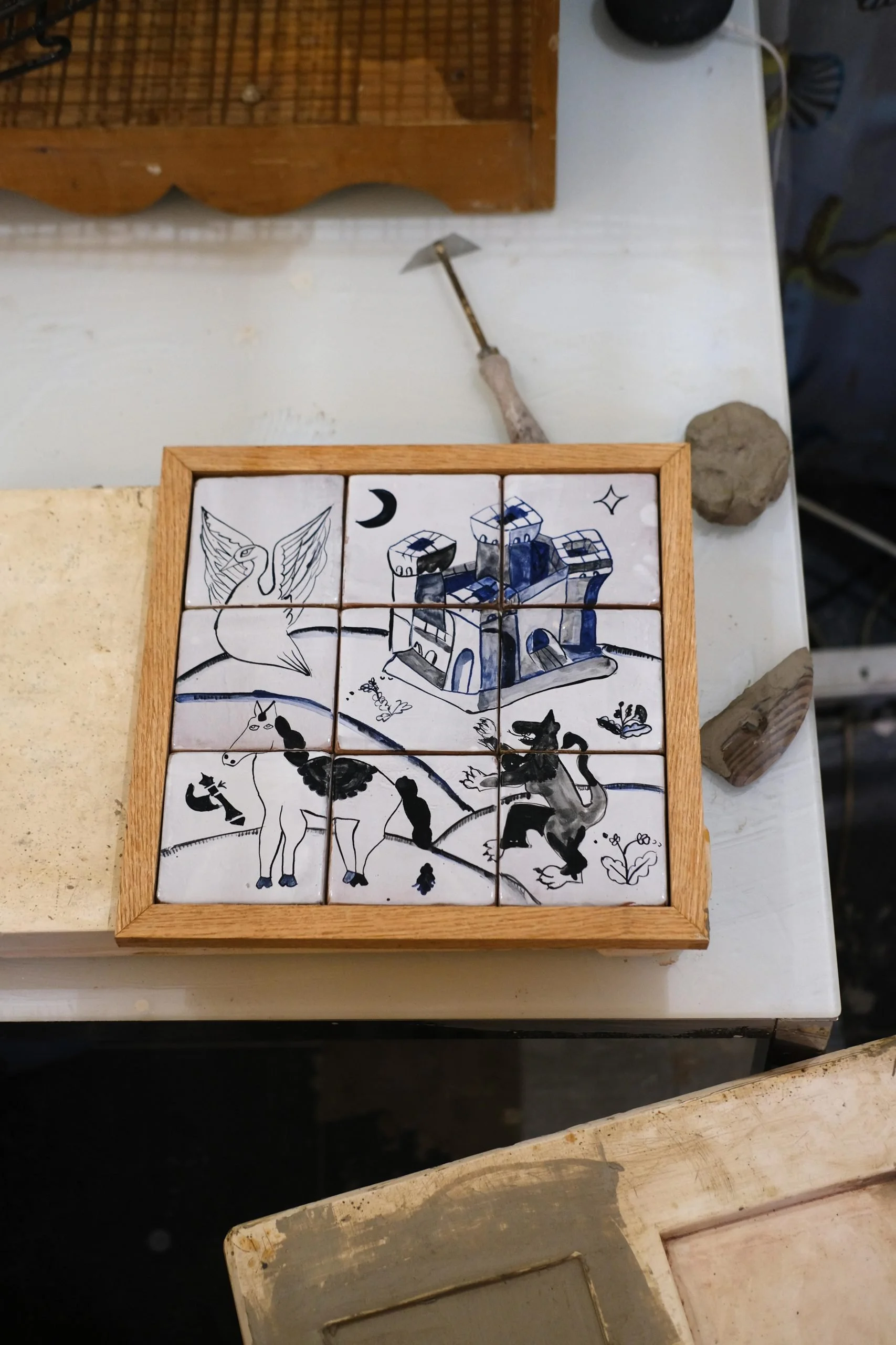 A wooden frame containing nine ceramic tiles with black and white illustrations of a castle, a swan, and animals including a dog, a cat, and a rabbit, arranged in a 3x3 grid.