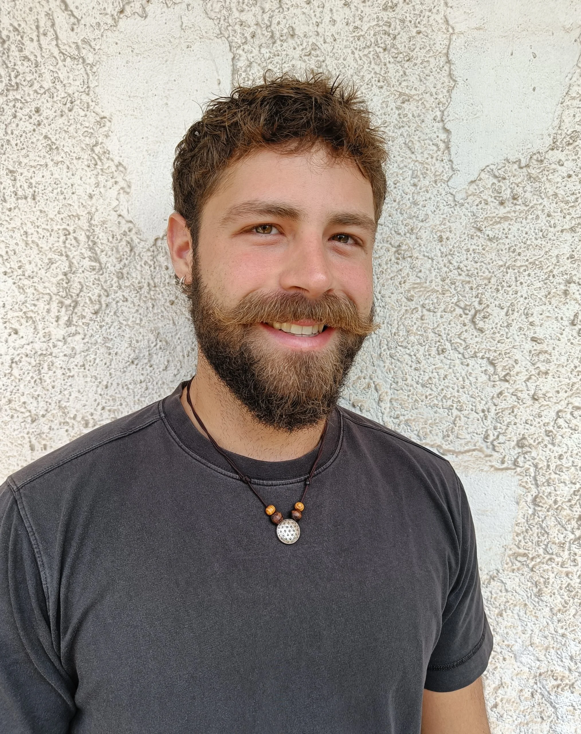 Close-up of a smiling man with a beard and mustache, wearing a black t-shirt and a beaded necklace with a decorative pendant, standing against a textured beige wall.