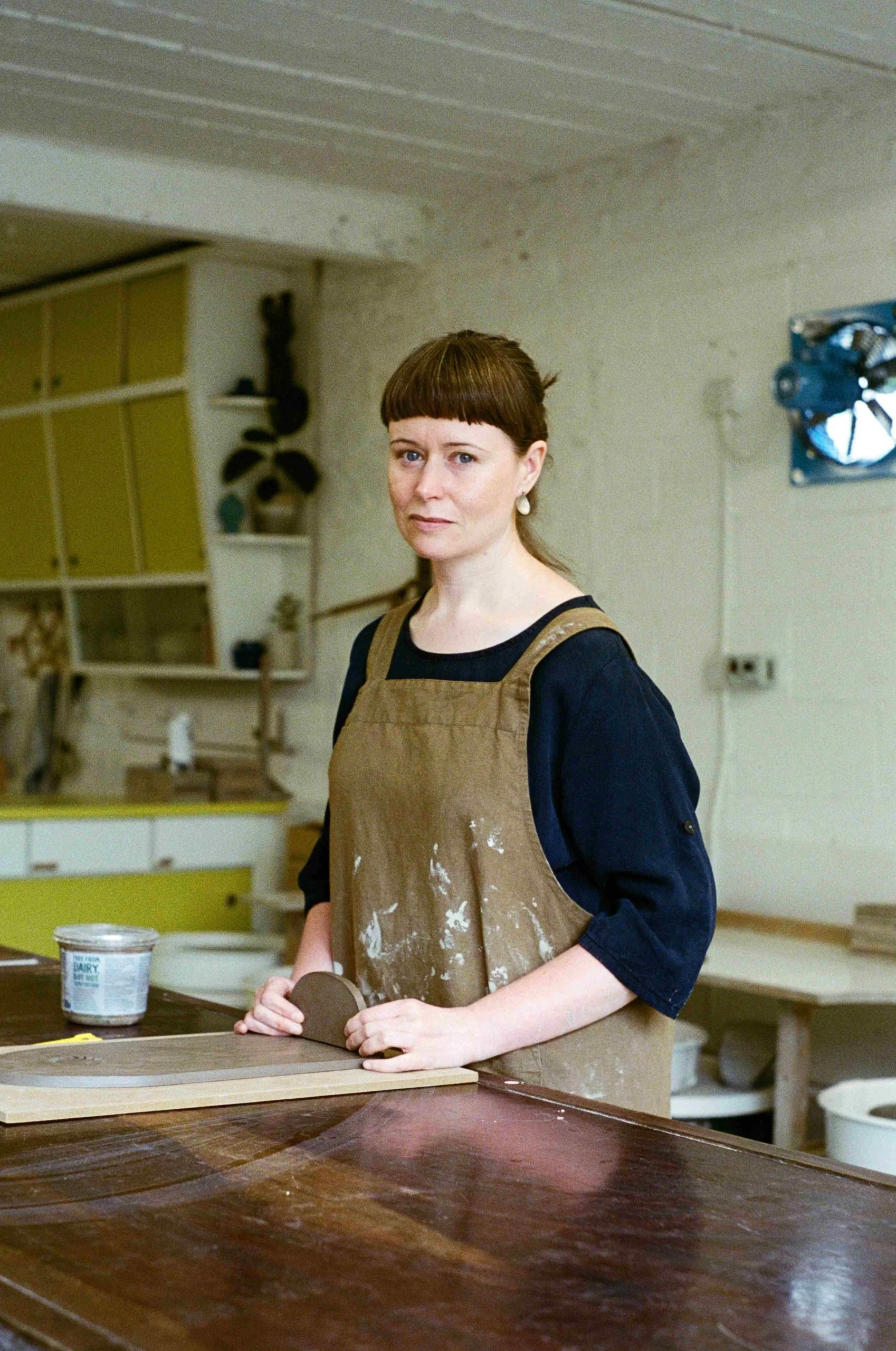 A woman with short brown hair and a brown apron standing in a light, airy pottery workshop. She is holding a rolling pin and rolling a large piece of clay.
