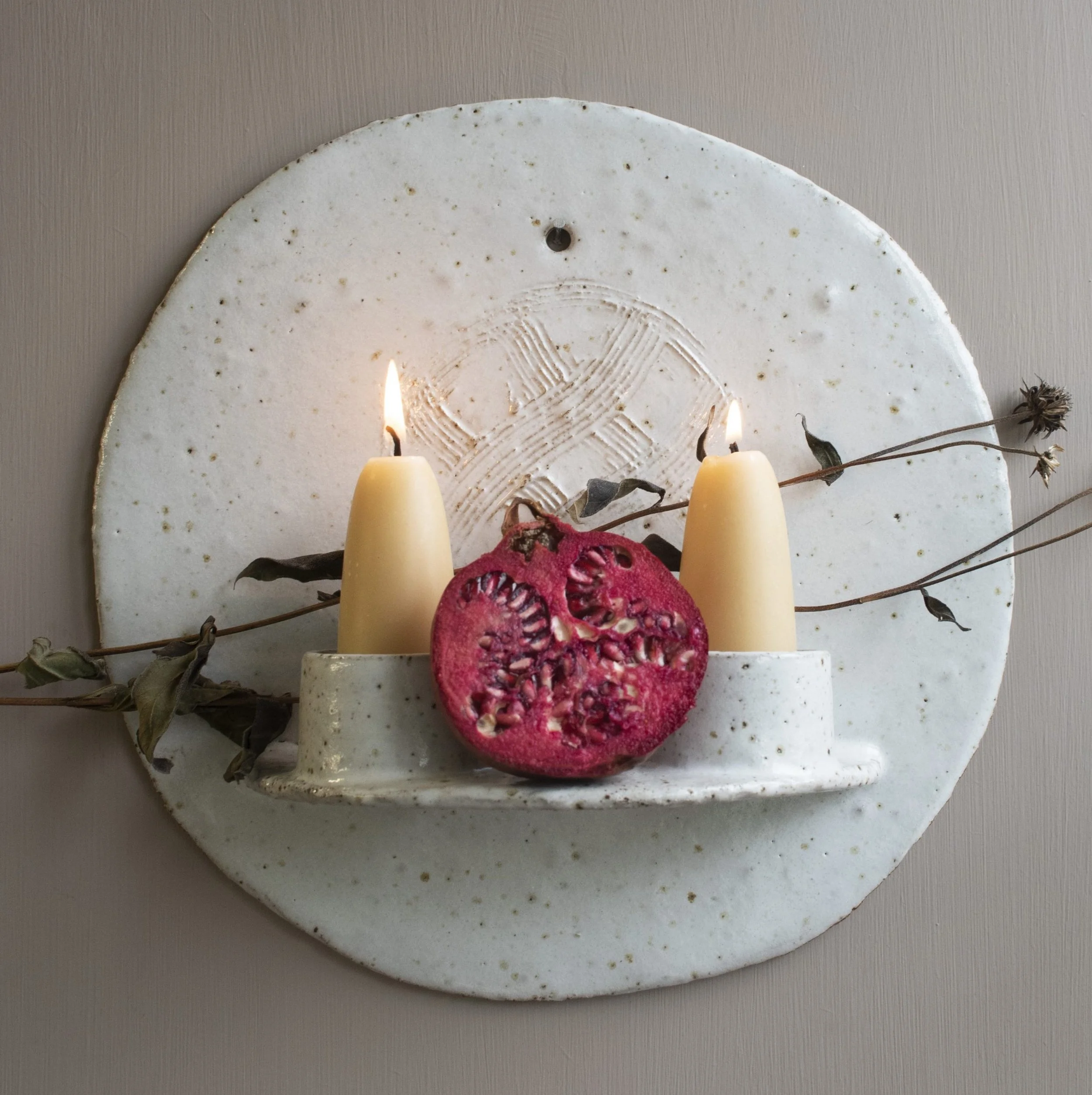 Close-up of a round white ceramic wall sconce with two white candles and dried plants, mounted on a light-colored wall.
