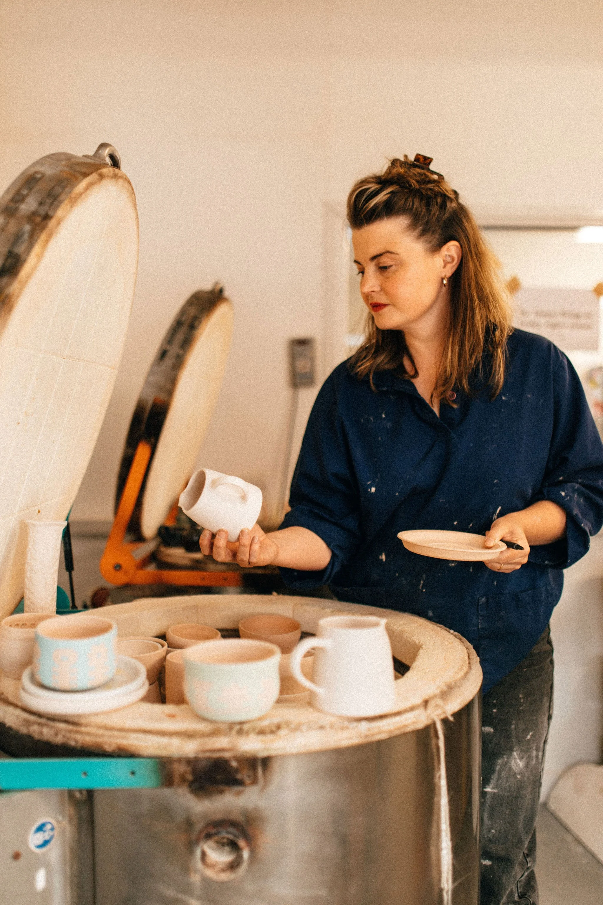 Woman working on pottery, unloading a finished large top loading kiln, holding a white ceramic piece in her hand.