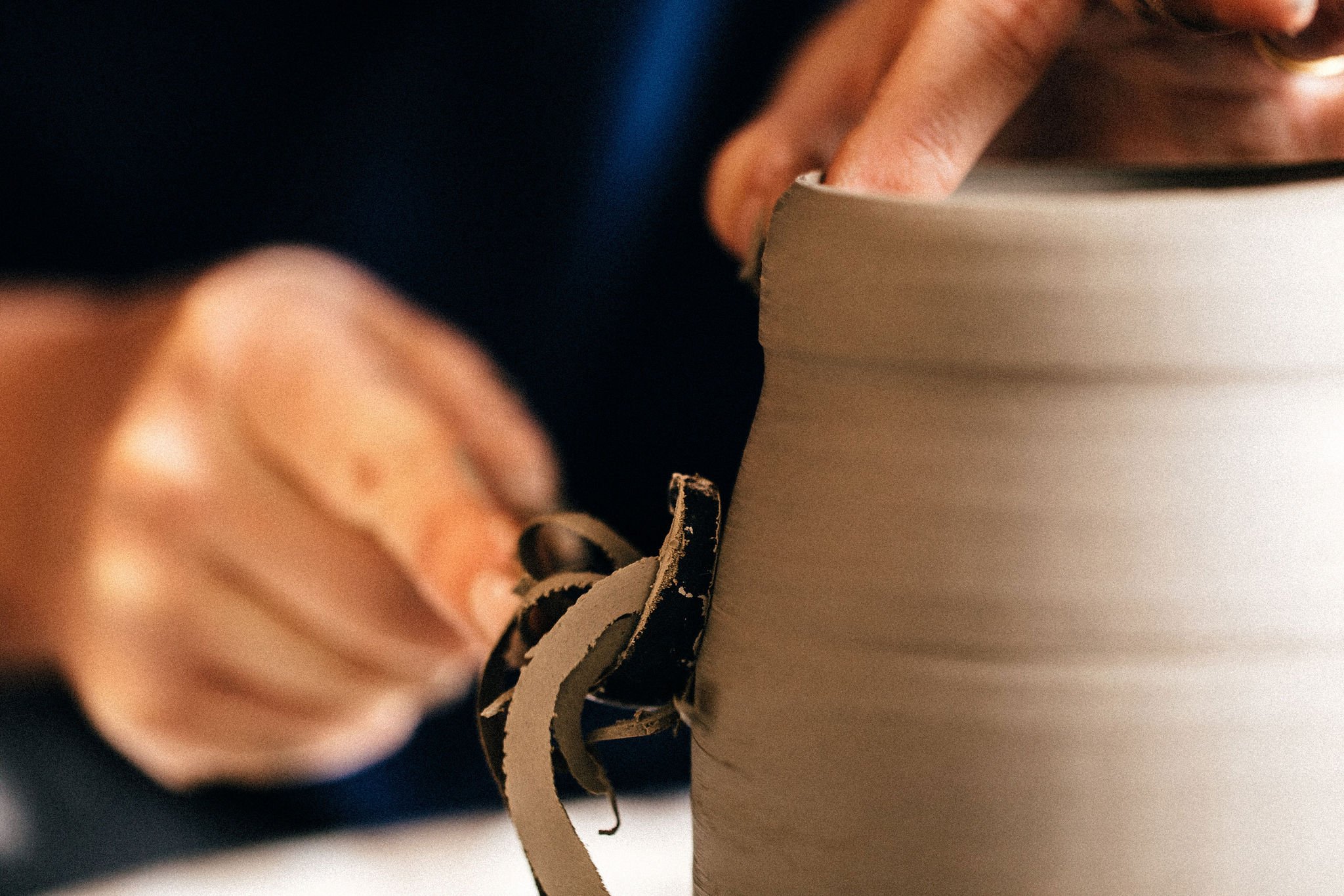 Close-up of a person's hands using a ceramic tool to trim excess clay from a wheel thrown pottery piece, using a pottery wheel.