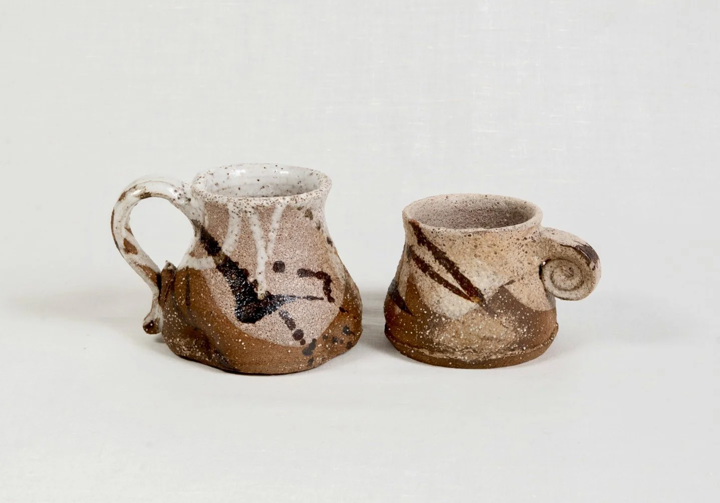 Two handmade pottery cups with brown and beige speckled glaze, featuring abstract black and brown painted designs, against a white background.