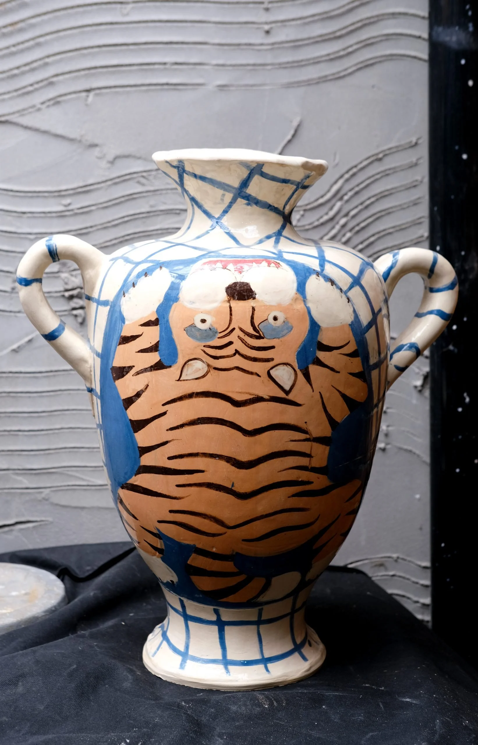 A ceramic vase decorated with a tiger face and striped pattern, featuring two handles and a flared top, set against a textured gray background.