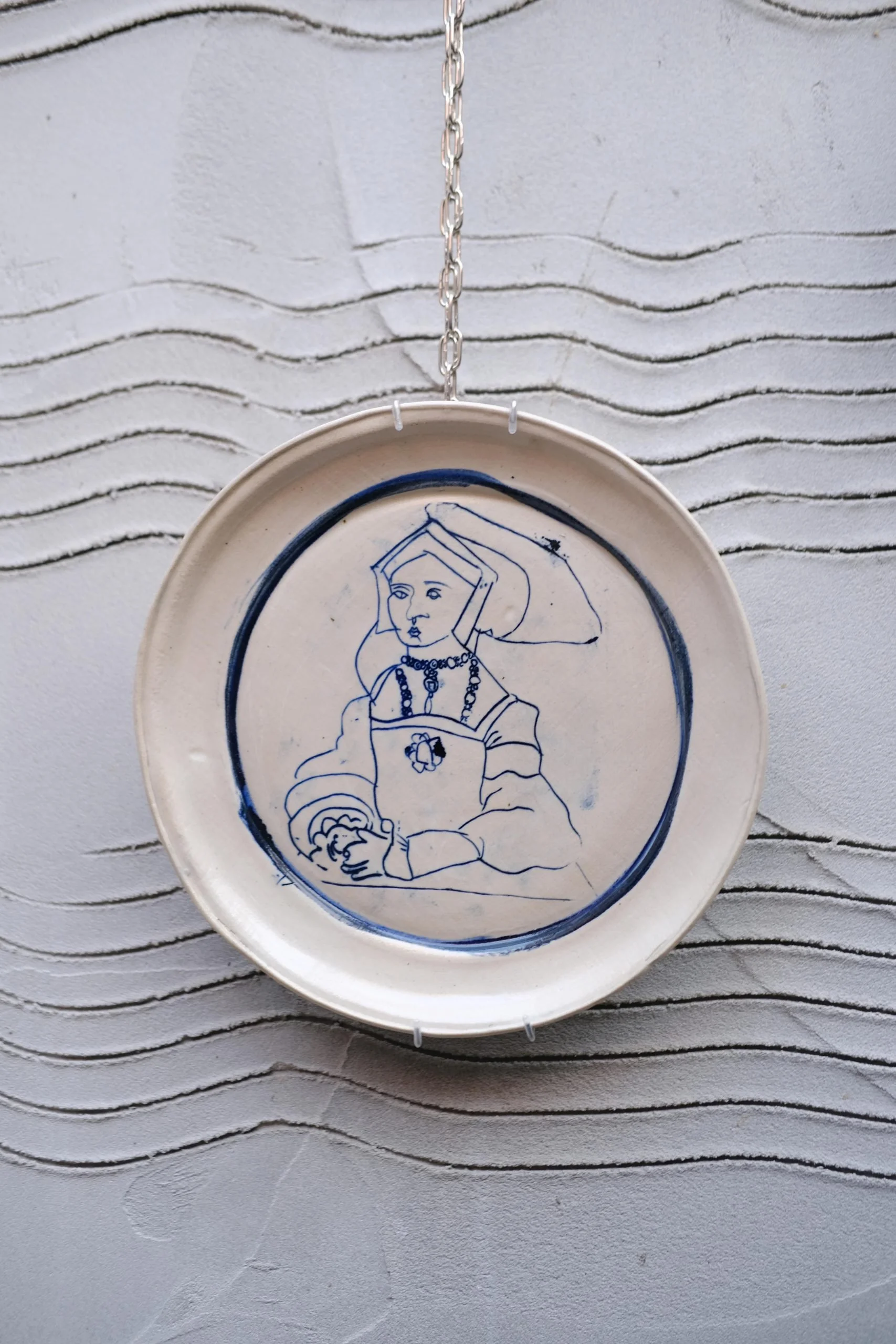 Decorative ceramic plate with a blue outline drawing of a woman holding a plate, hanging from a metal chain on a textured wall.