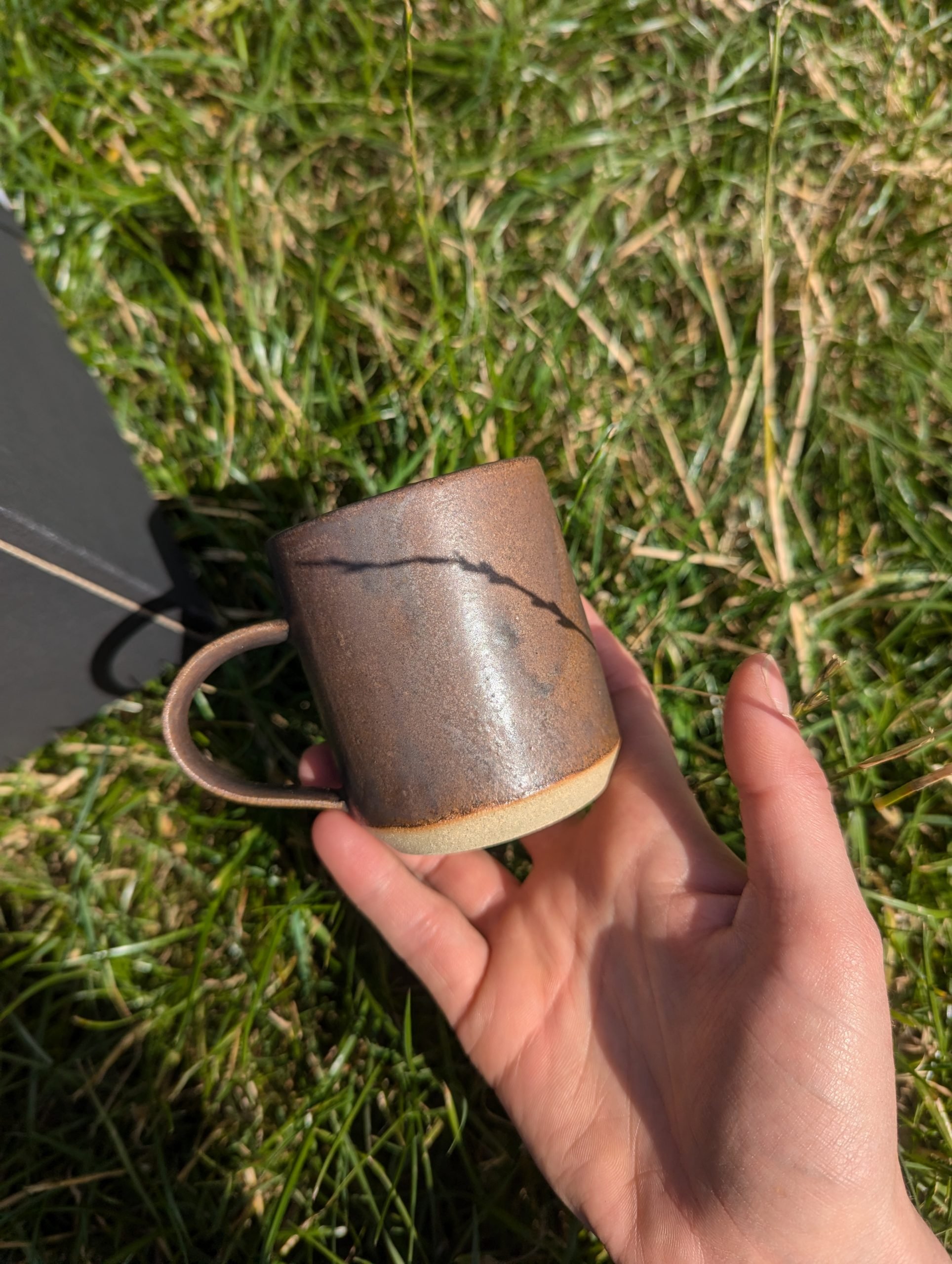 Hand holding a rust coloured, metallic glazed mug against a background of green grass.
