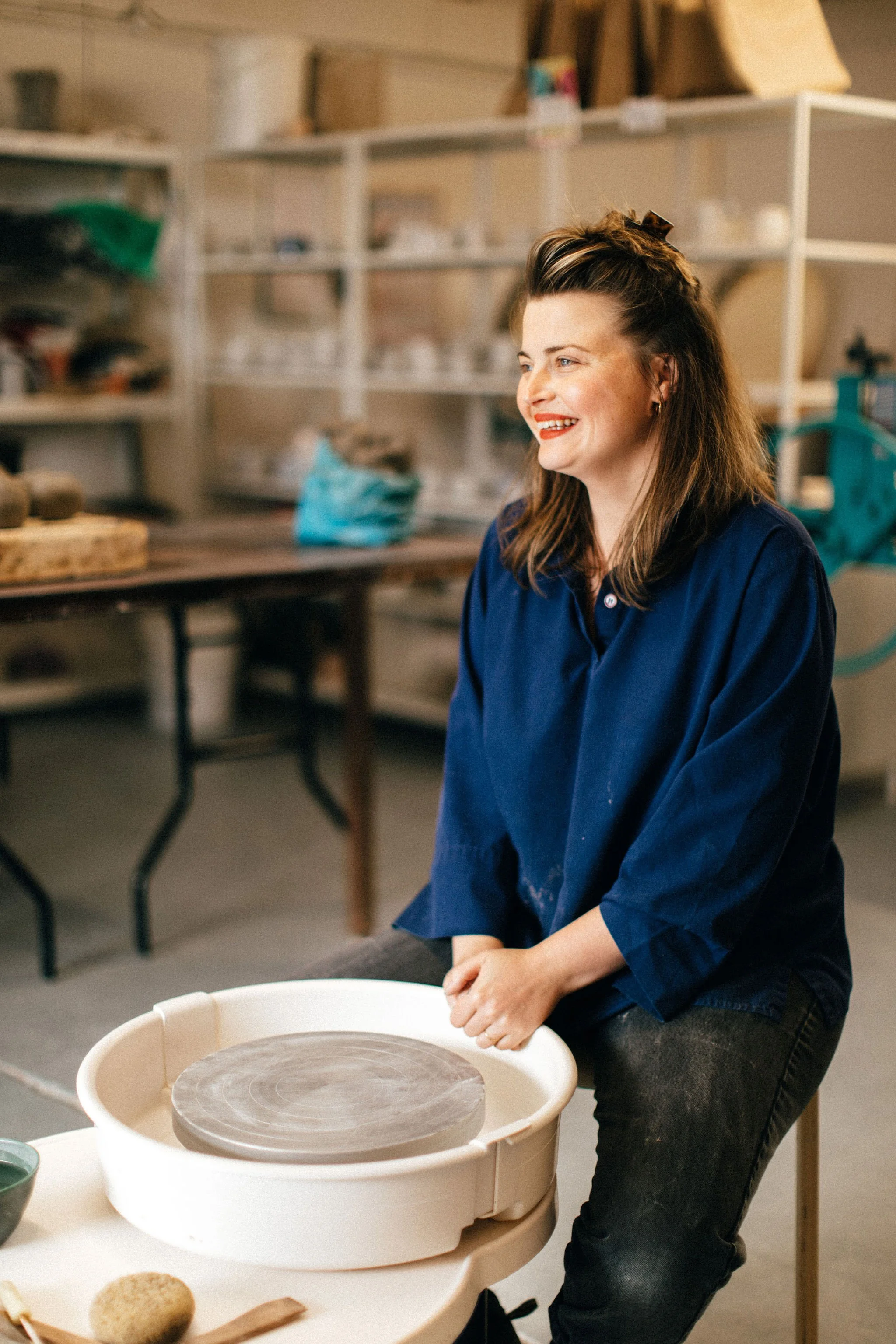 A woman with brown hair wearing a blue shirt and black pants, smiling as she works with a pottery wheel in a ceramics studio.