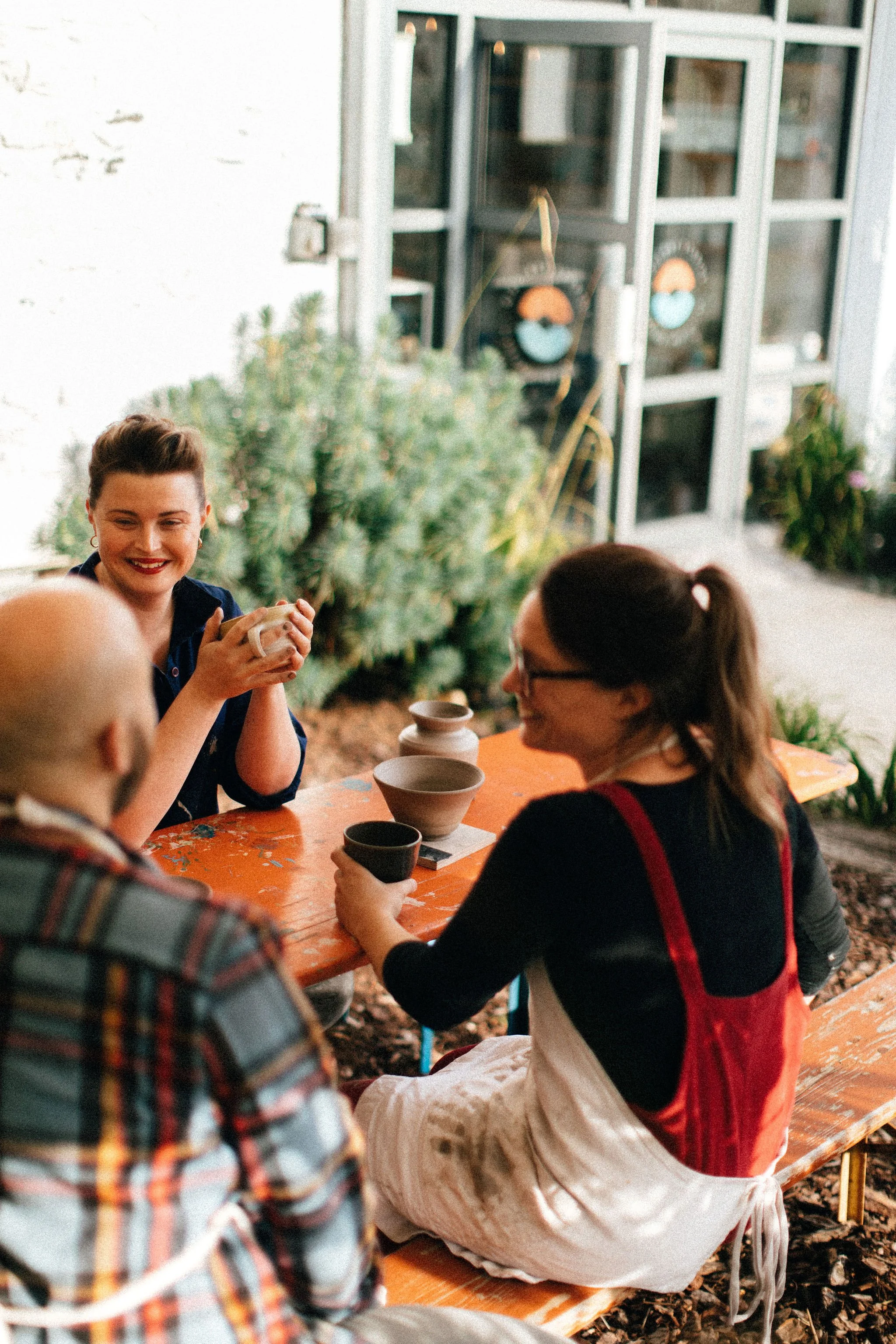 Group of people socializing outdoors at a wooden picnic table, with tea cups and a plant in the background.