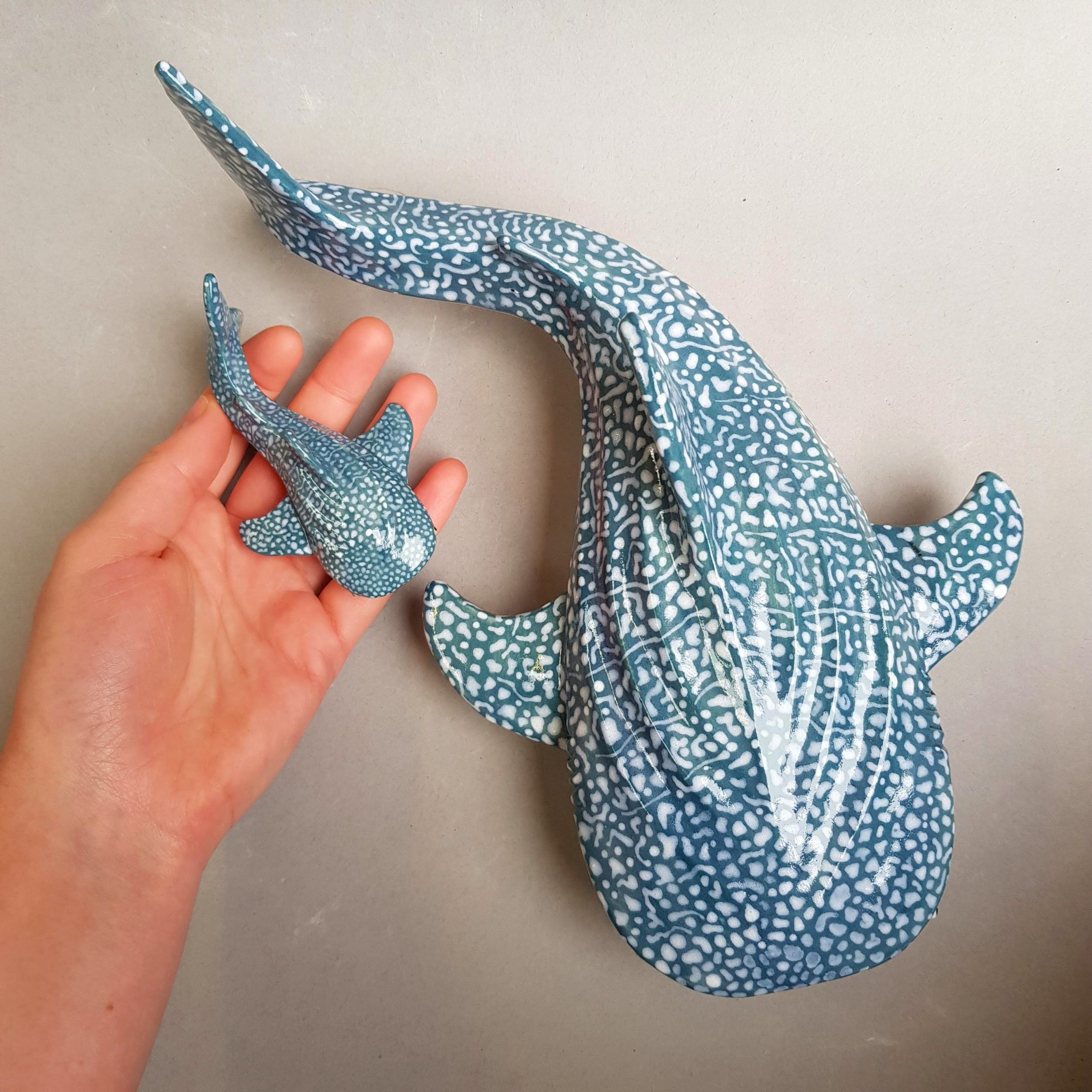 A person's hand holding a small, blue animal-shaped ceramic figurine with a white dotted pattern, and a larger, matching animal-shaped figurine in the background, both on a flat, light-colored surface.