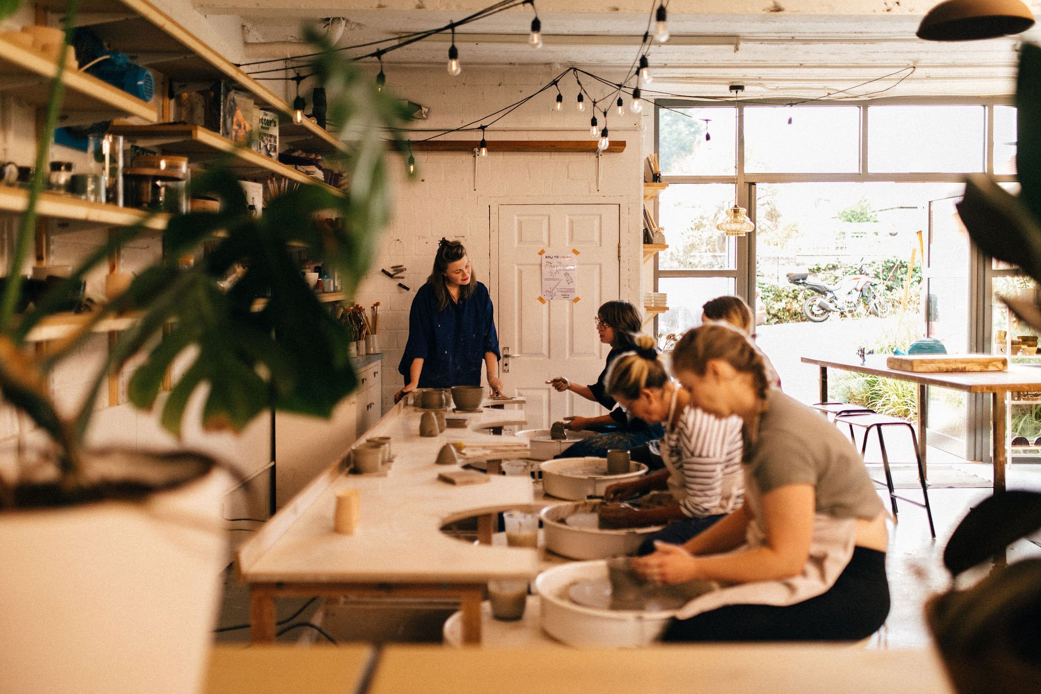 People participating in a ceramics class in a bright, airy studio with large windows, wooden shelves, and string lights.