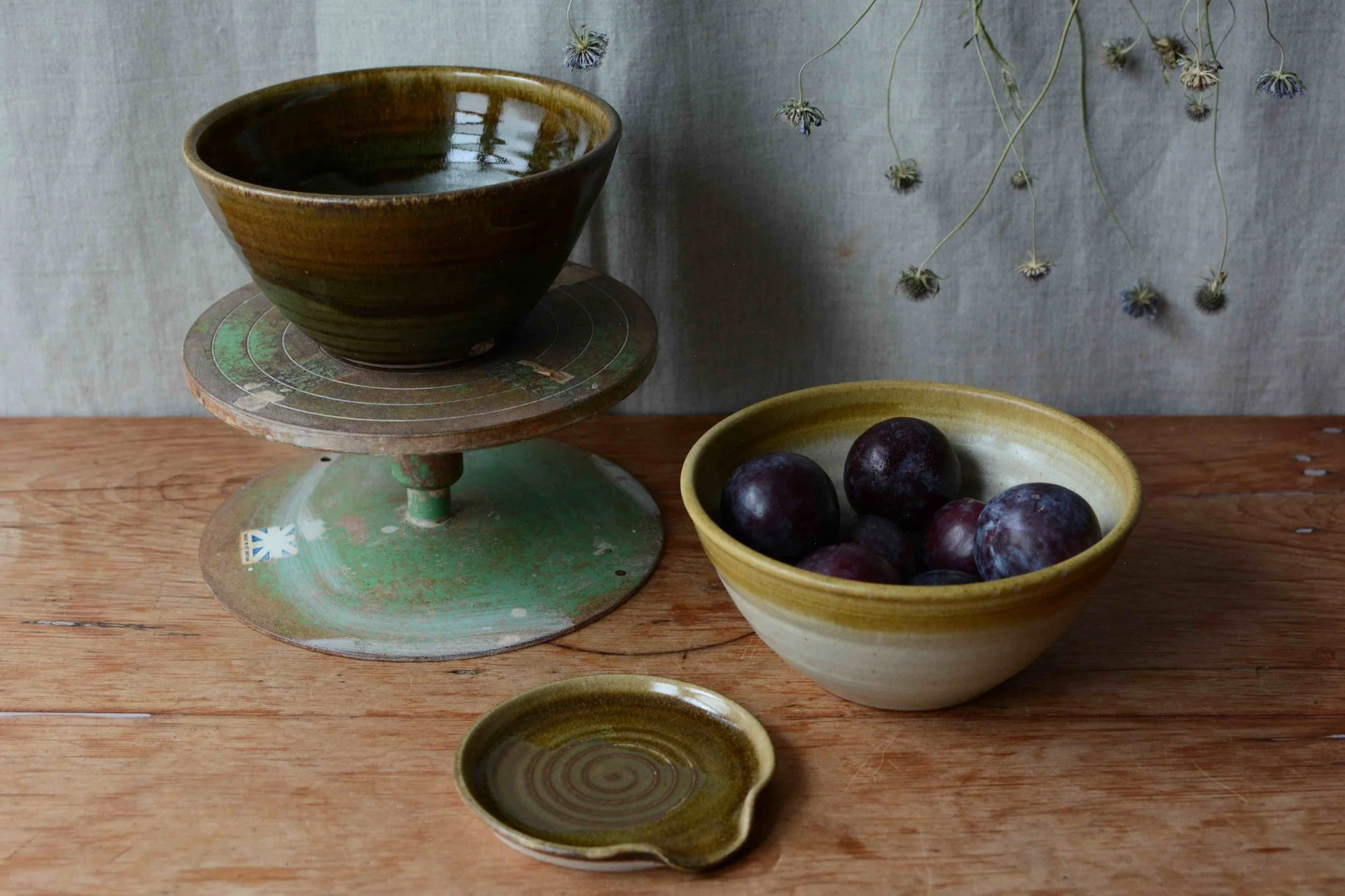 A rustic wooden table holds a green ceramic bowl filled with dark grapes, a small green and brown ceramic pitcher, a vintage green and wood cake stand with a brown ceramic bowl, and a small bowl with a spiral design. In the background, a gray fabric backdrop and dried flowers hang upside down.
