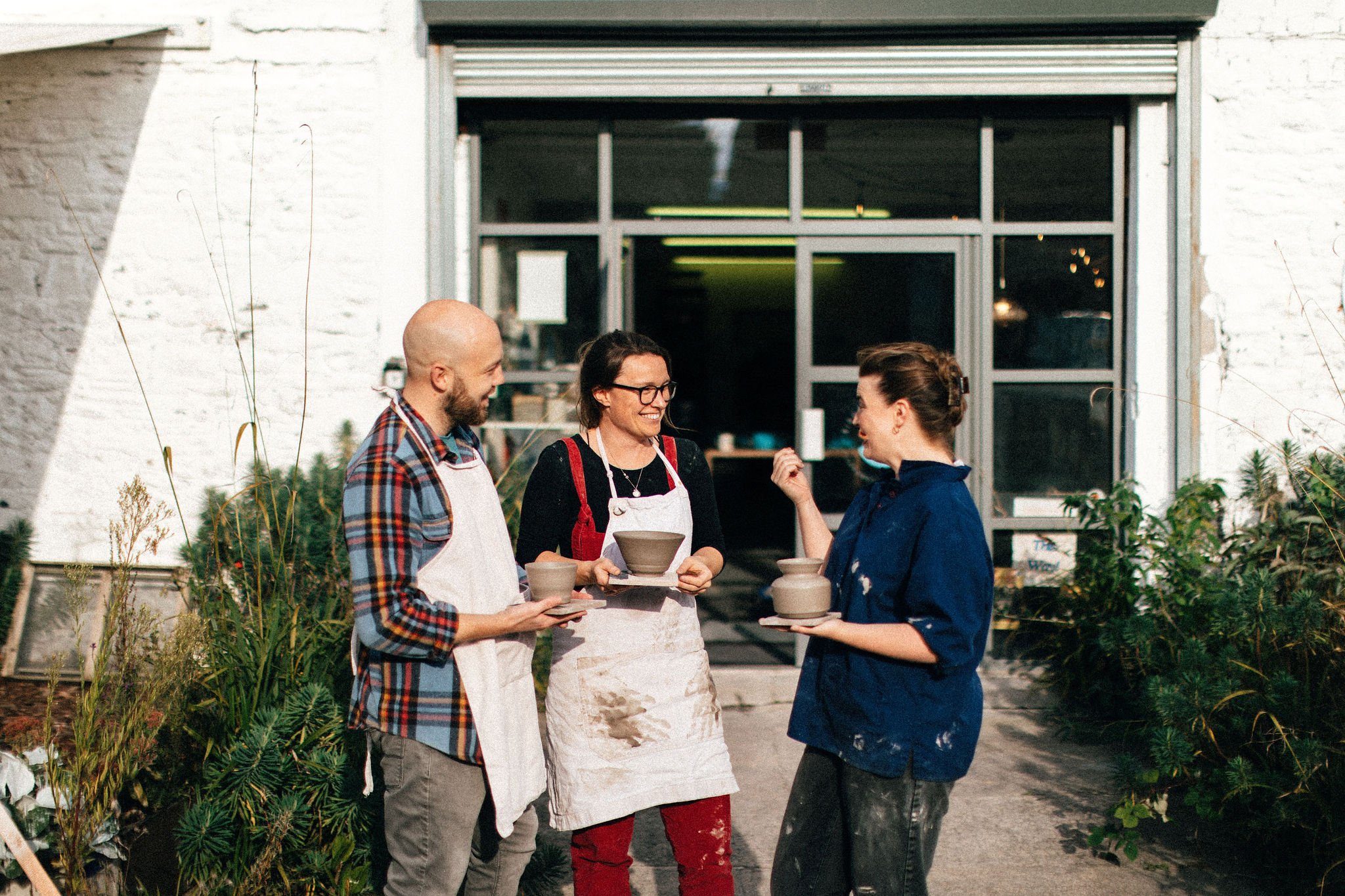 Three people smiling and talking outside of a building, with two of them holding clay pots. They appear to be at a pottery workshop, with plants nearby.