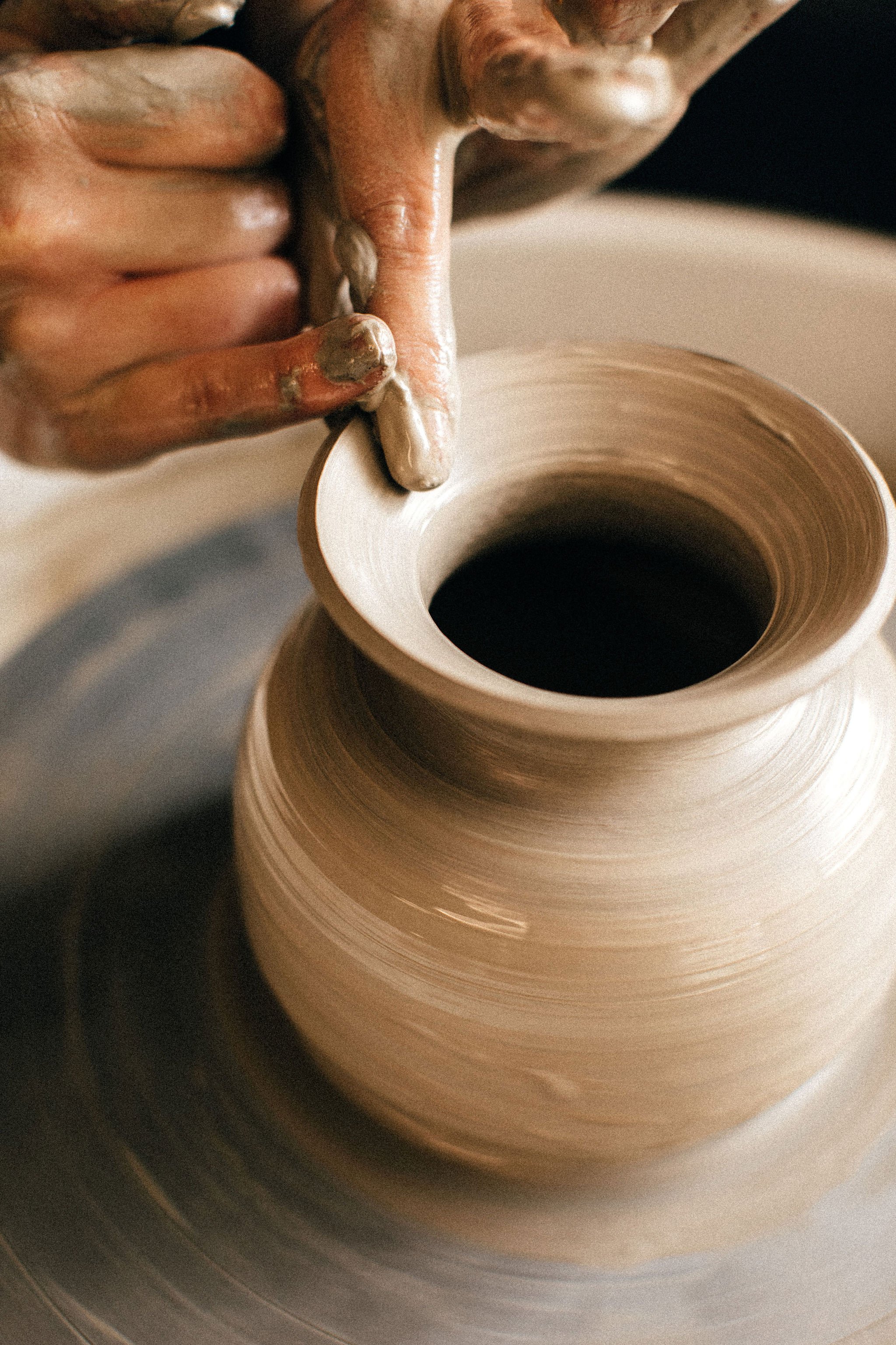 Close-up of a person shaping a clay pottery wheel, hands covered in wet clay, with a partially formed ceramic vessel in progress.