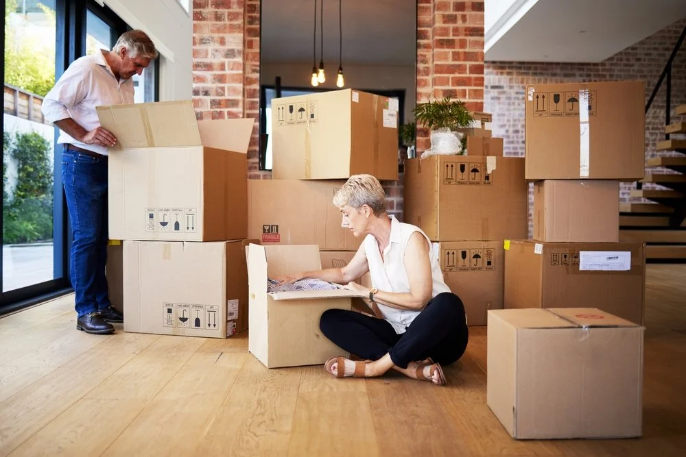 Two people packing or unpacking moving boxes inside a home with wood floors and brick walls. One person is sitting on the floor looking at an open box, and the other is standing and looking into a larger box.