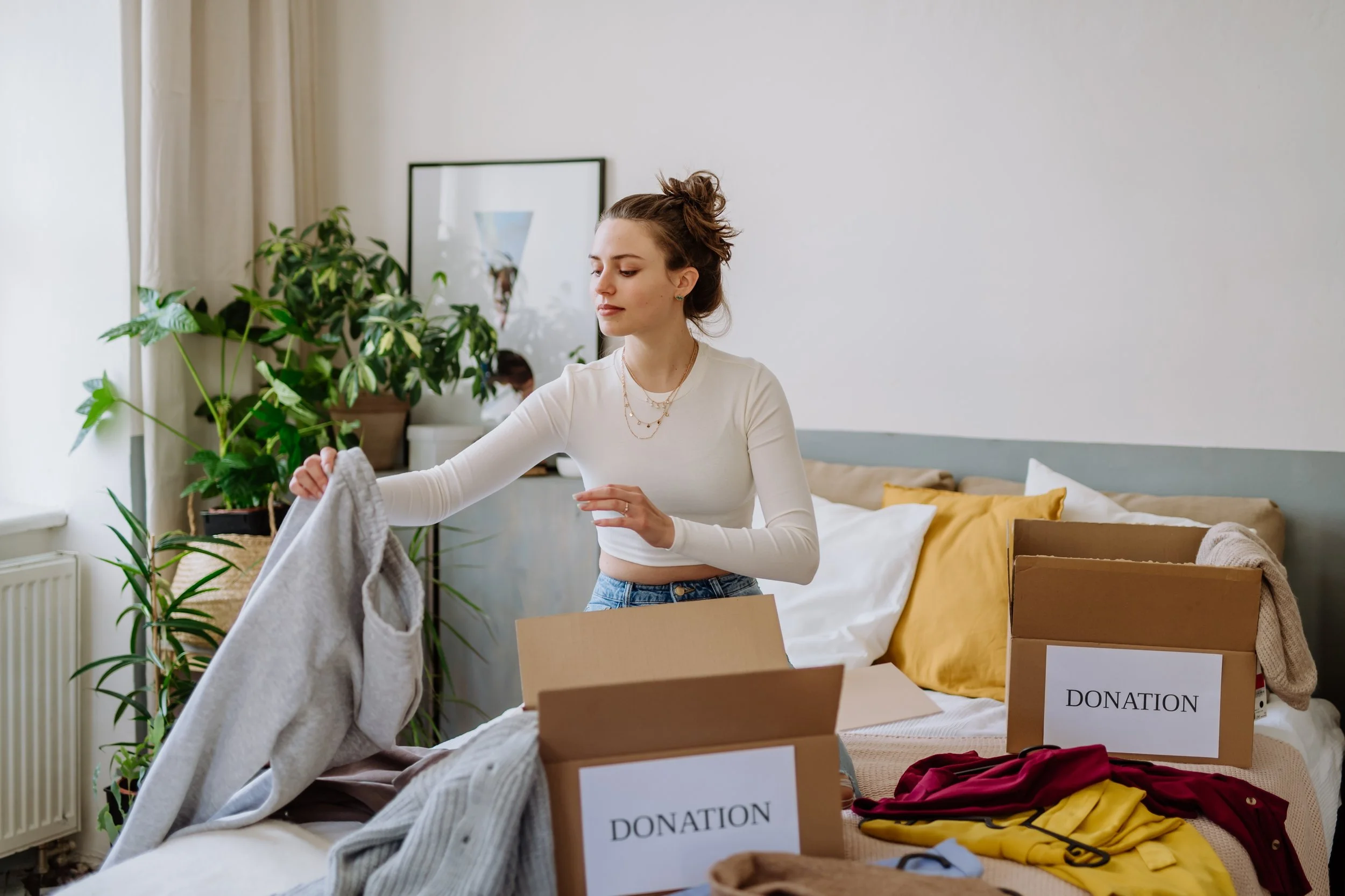 A young woman folding clothes in a room with donation boxes on a bed and plants in the background.