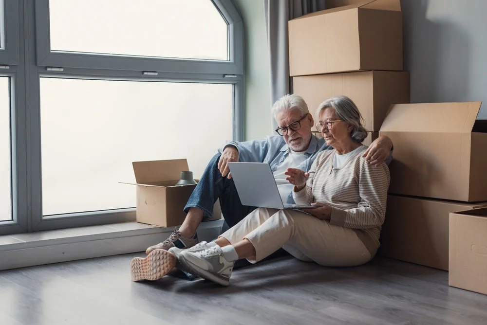 An elderly couple sitting on the floor of a new apartment with cardboard moving boxes, looking at a tablet together.