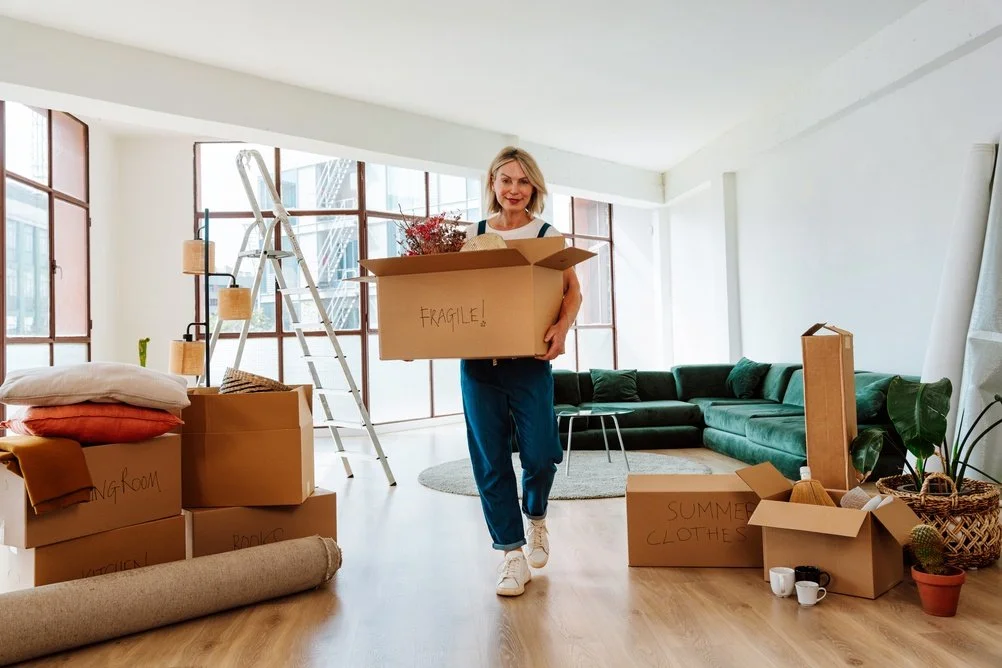 Woman moving boxes filled with household items in a bright living room, with a ladder, green sectional sofa, and large windows.