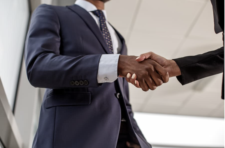 Two people shaking hands, one in a navy suit and the other in a black suit, in a professional setting.