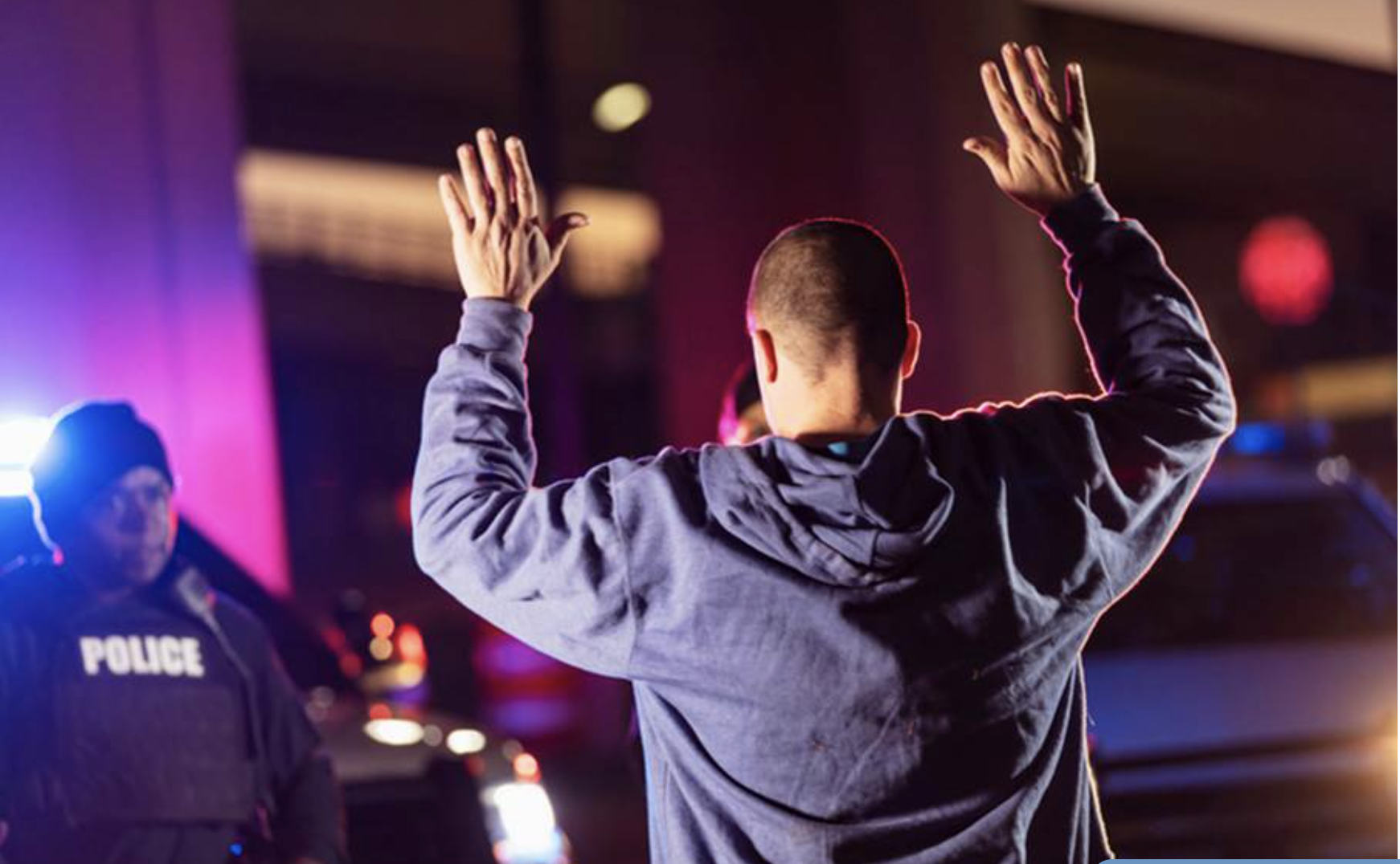 A man with a shaved head in a gray hoodie with arms raised being detained by police at night, with police officers and police cars visible in the background