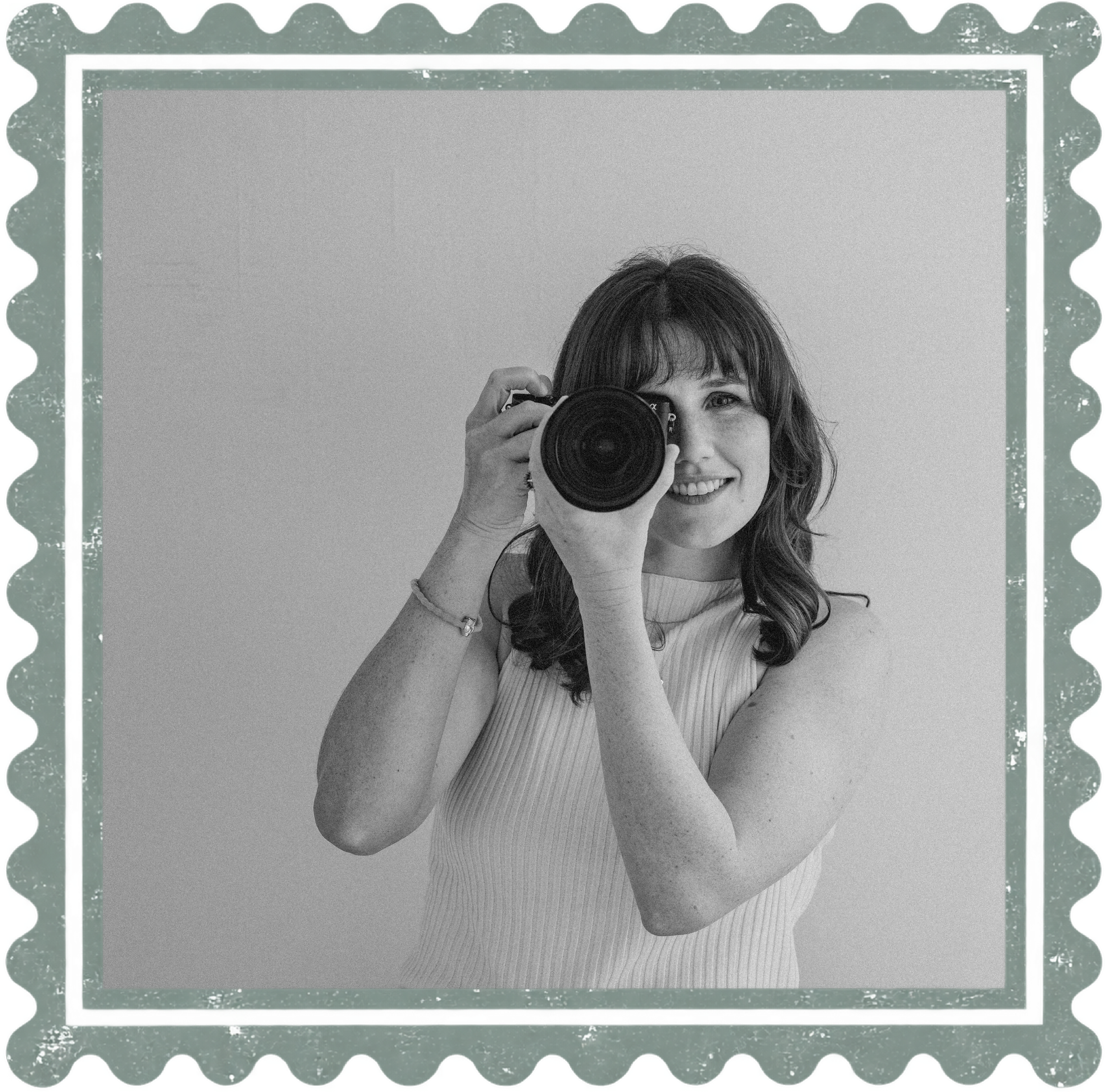 A woman with long hair taking a selfie using a camera, smiling, with a plain wall in the background and a decorative green border around the image.