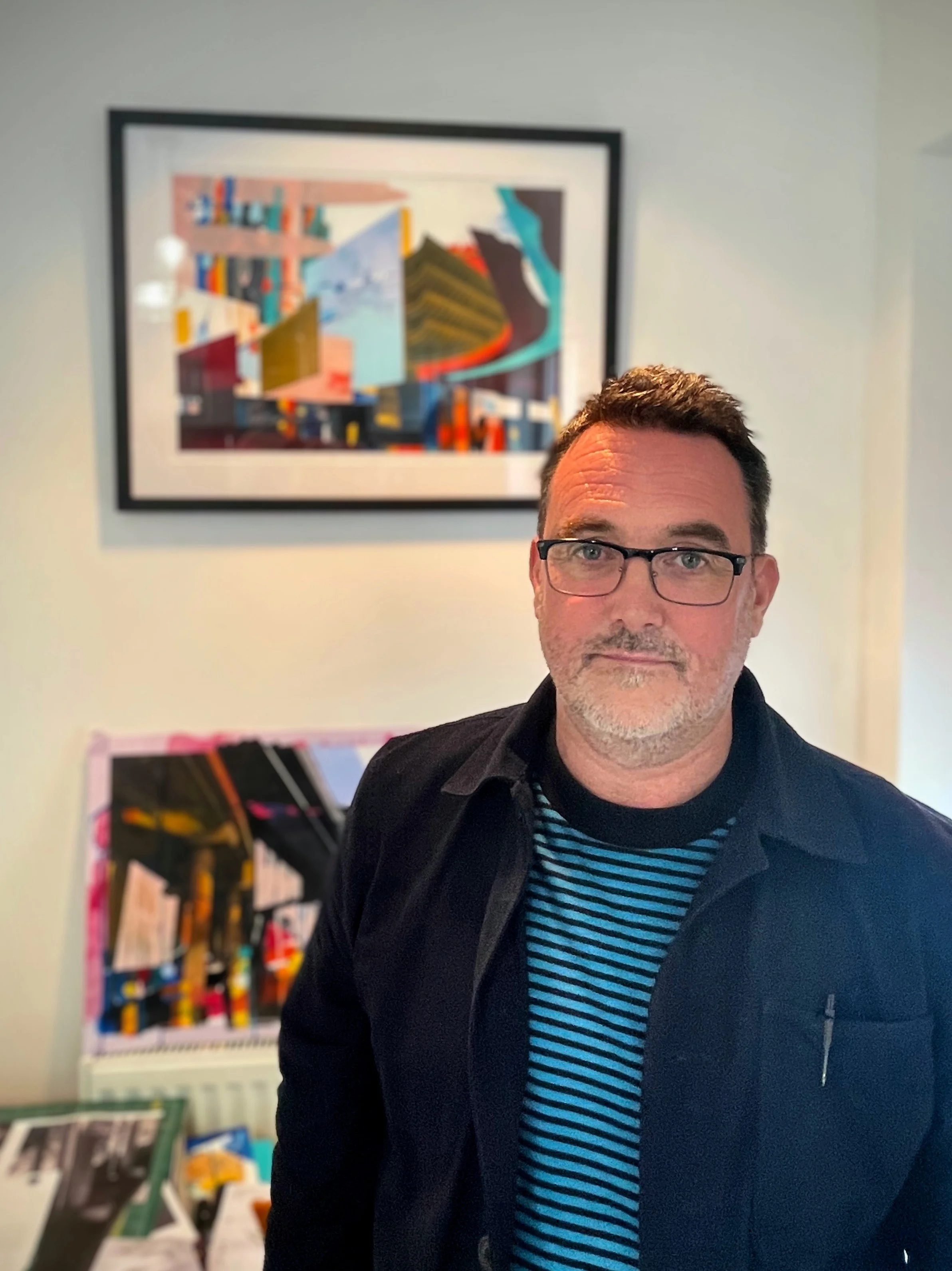 A man with glasses and a beard wearing a black jacket and a blue-striped shirt, standing in front of colorful artwork on a white wall.