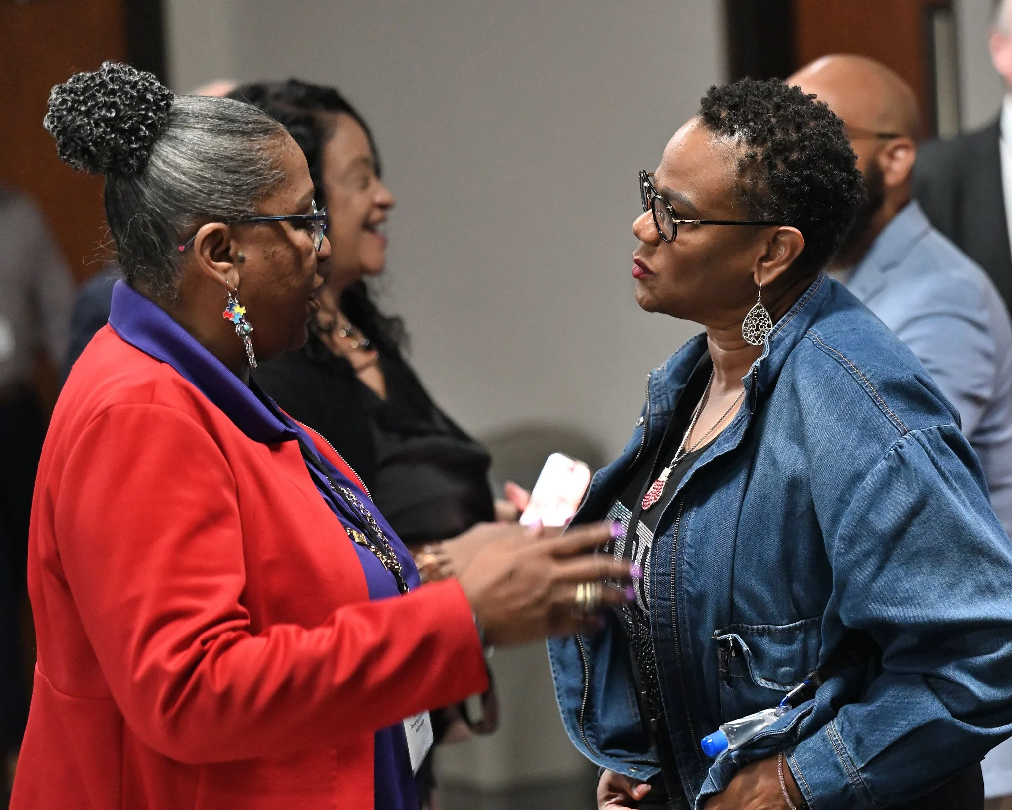 Two women engaged in conversation, facing each other, in what appears to be a professional or formal setting, with other people in the background.